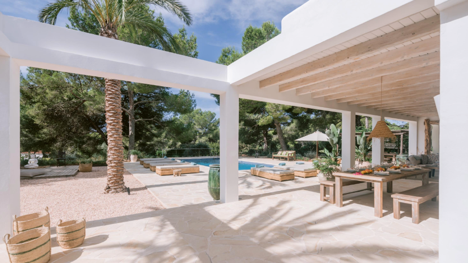 An outdoor patio with a dining table, benches, and a view of a backyard with a swimming pool, lounge chairs, an umbrella, lush greenery, and palm trees under a partly cloudy sky.