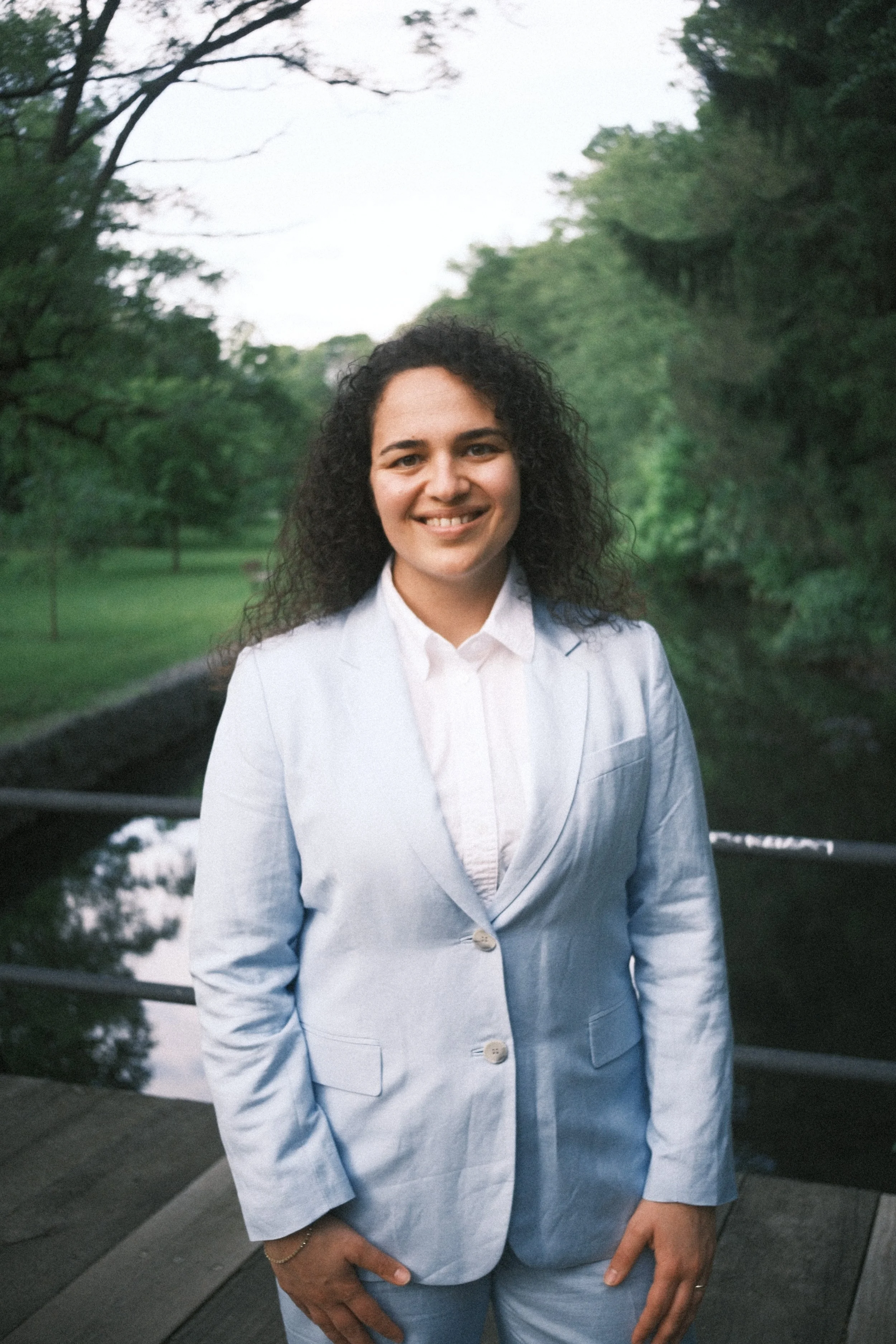 A woman with curly dark hair, smiling, wearing a light blue suit and white shirt, standing outdoors near a river and trees, during daytime.