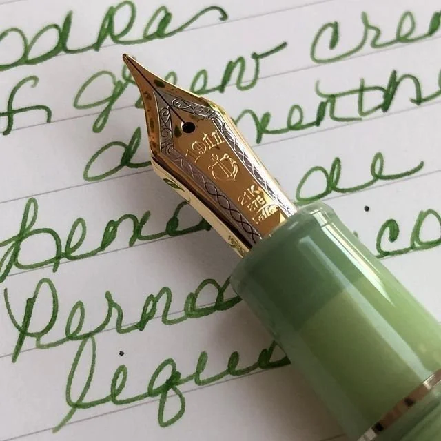 Close-up of a fountain pen with a gold nib writing green ink cursive on white lined paper.