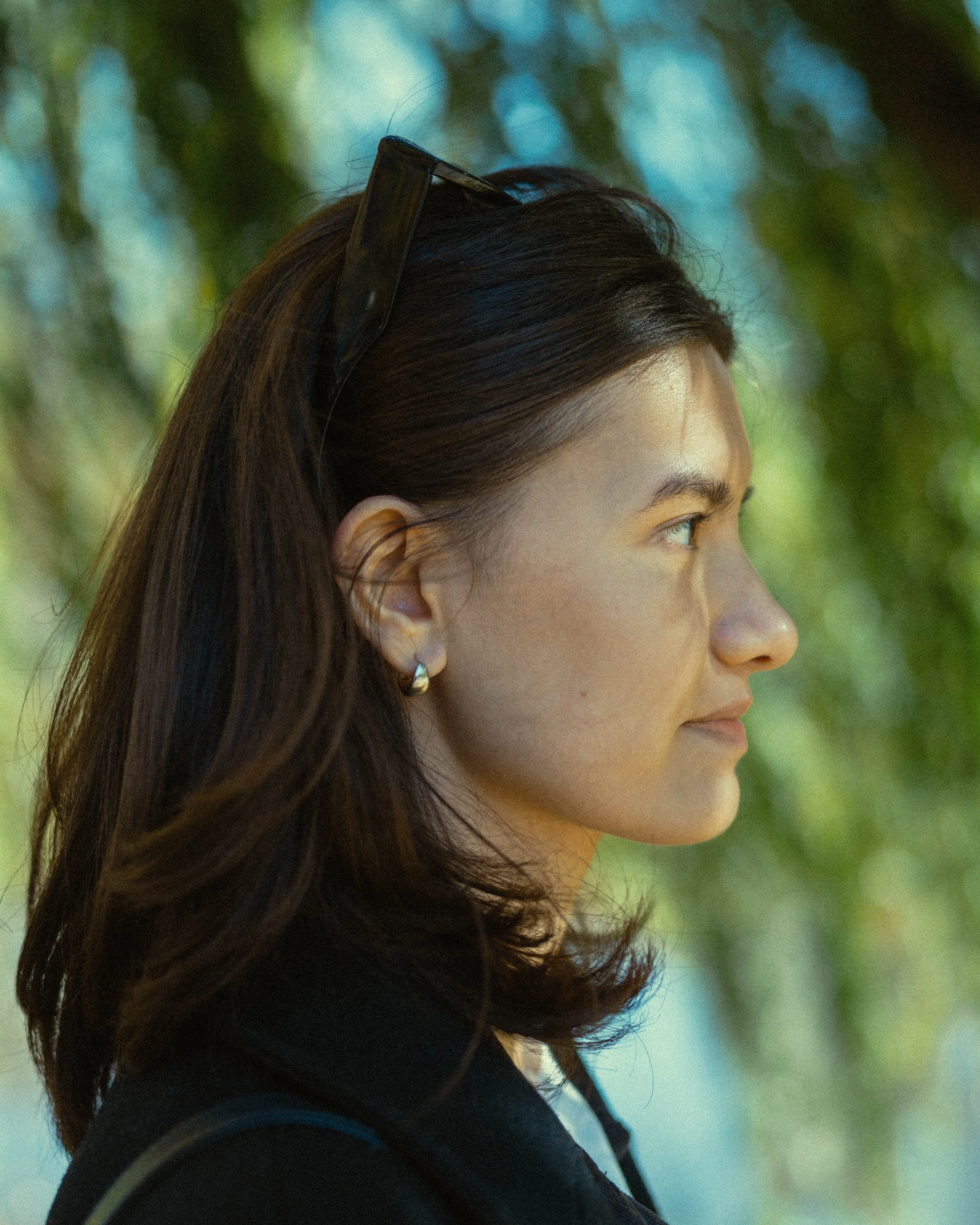 Close-up side profile of a woman with long brown hair, wearing sunglasses on her head, earrings, and a dark jacket, outdoors with green and blue blurred background.