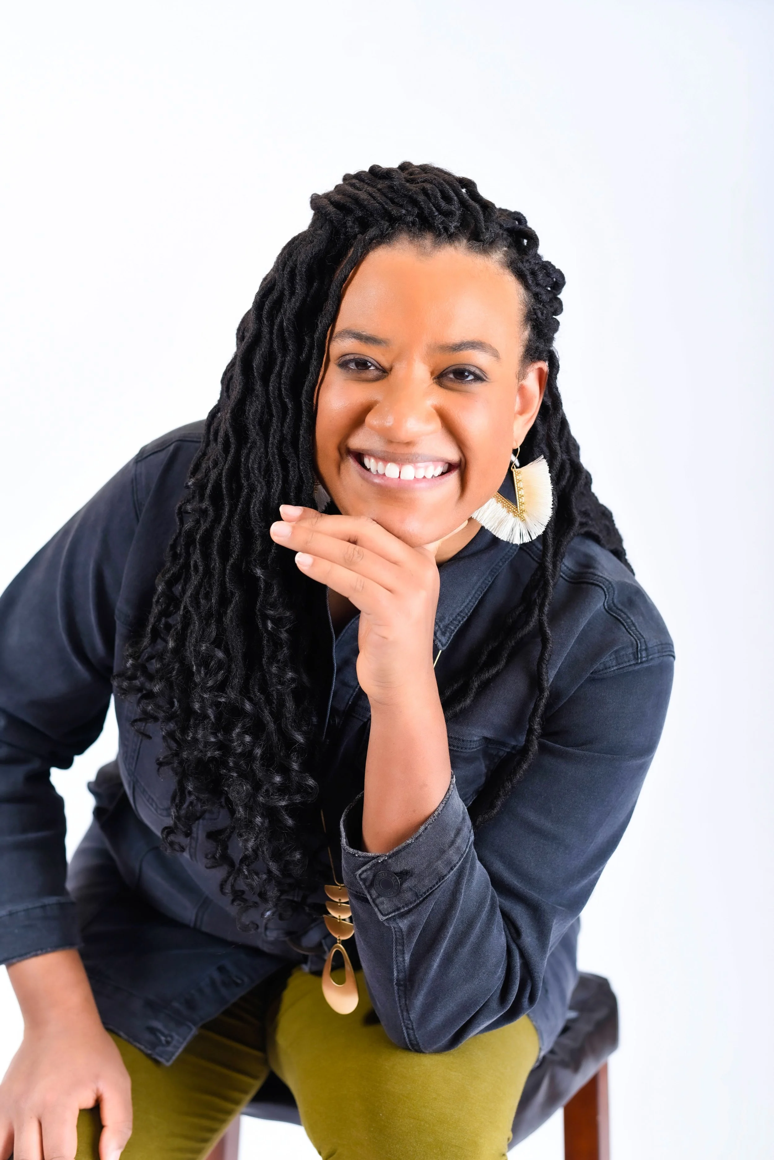 A woman with long curly black hair and earrings, smiling and resting her chin on her hand, sitting on a chair against a plain white background.