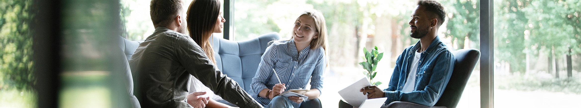Four young adults having a conversation in a bright, modern room with large windows and greenery outside.