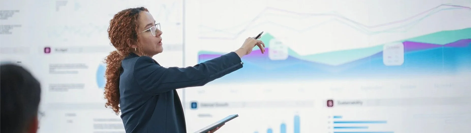 Woman with curly red hair and glasses pointing at a large screen displaying graphs and charts during a presentation.