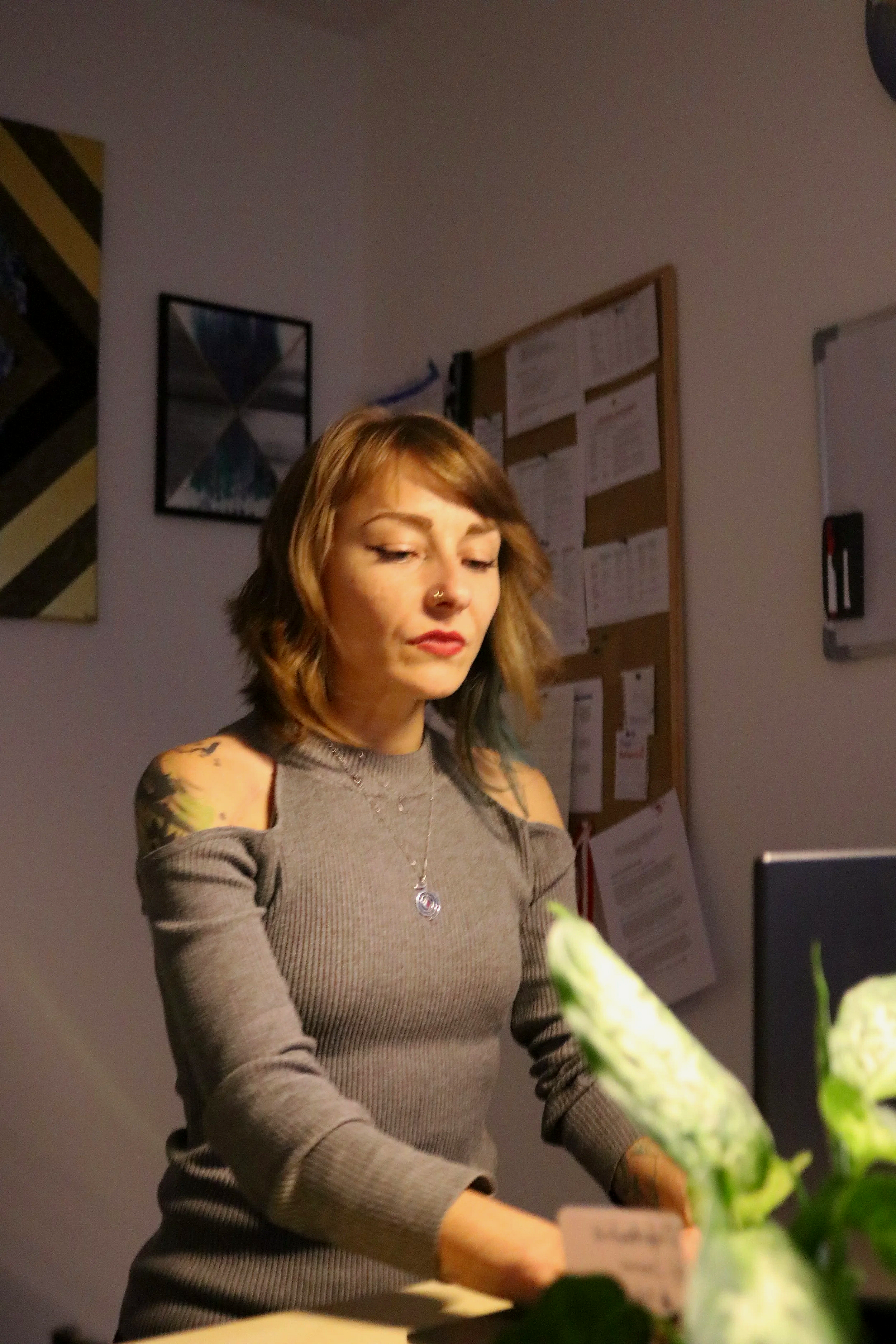 A young woman with short brown hair, wearing a gray cold-shoulder top, standing in front of a bulletin board and some artwork on the wall, looking down at what she is doing.
