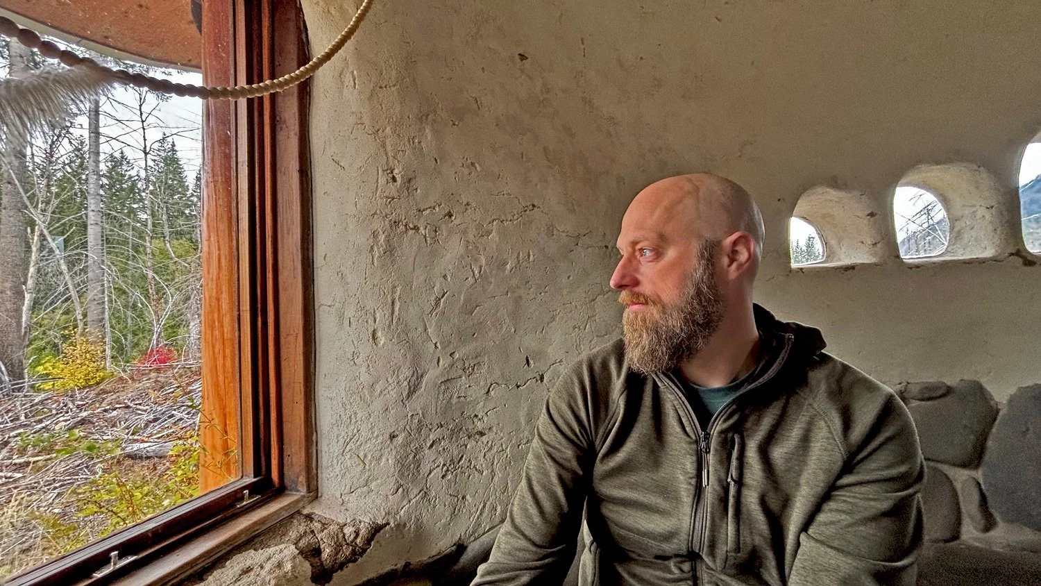 Man with a beard sitting indoors, looking out the window at the trees outside.