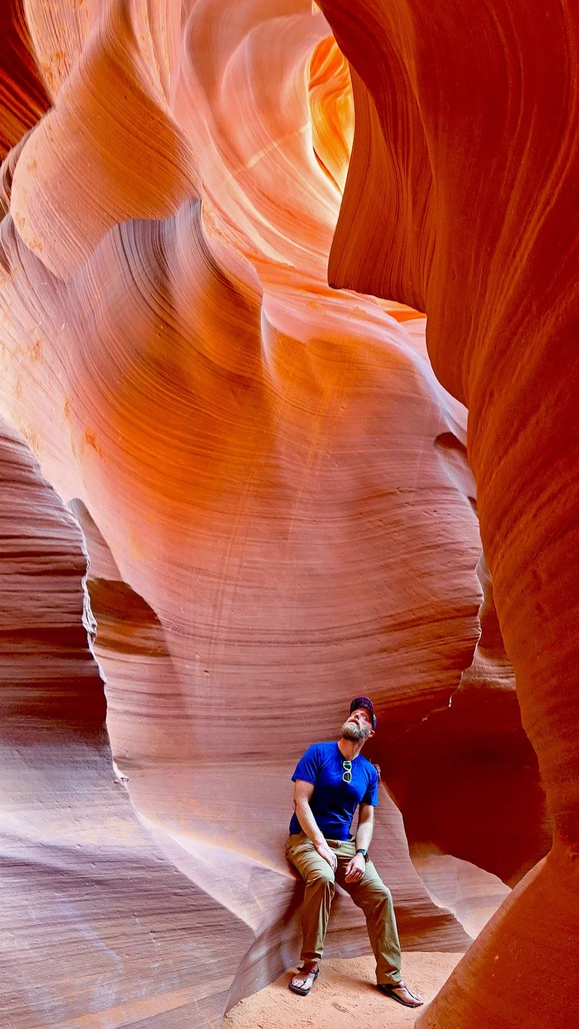A man wearing a blue shirt, tan pants, and flip-flops sitting in a narrow, winding slot canyon with smooth, layered sandstone walls in shades of orange and red.