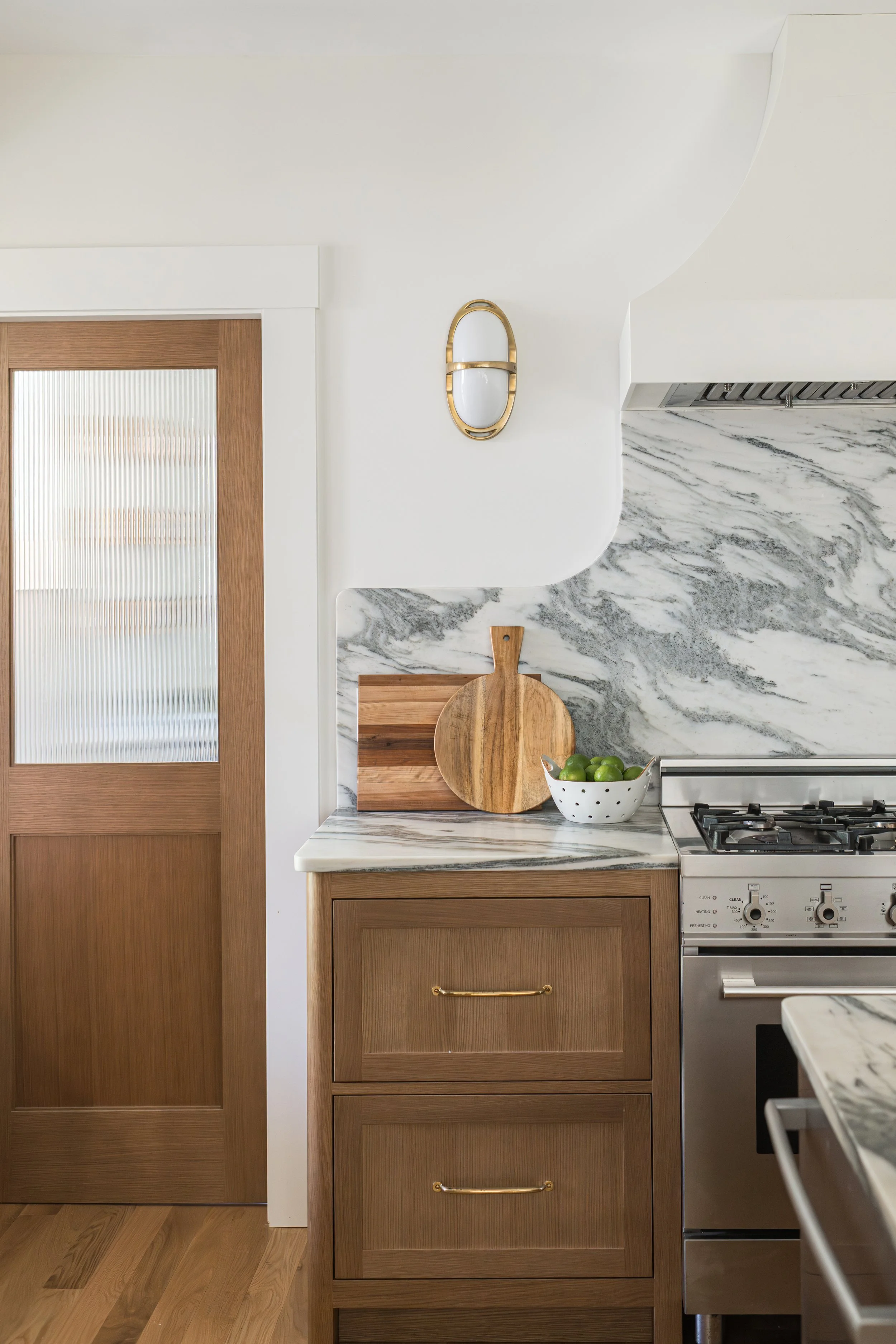 A kitchen scene featuring a wooden cabinet, a marble countertop with a wooden cutting board and a bowl of green apples, a stove, and a wall with a white and gray marble backsplash. A decorative wall sconce with gold accents is mounted on the wall.