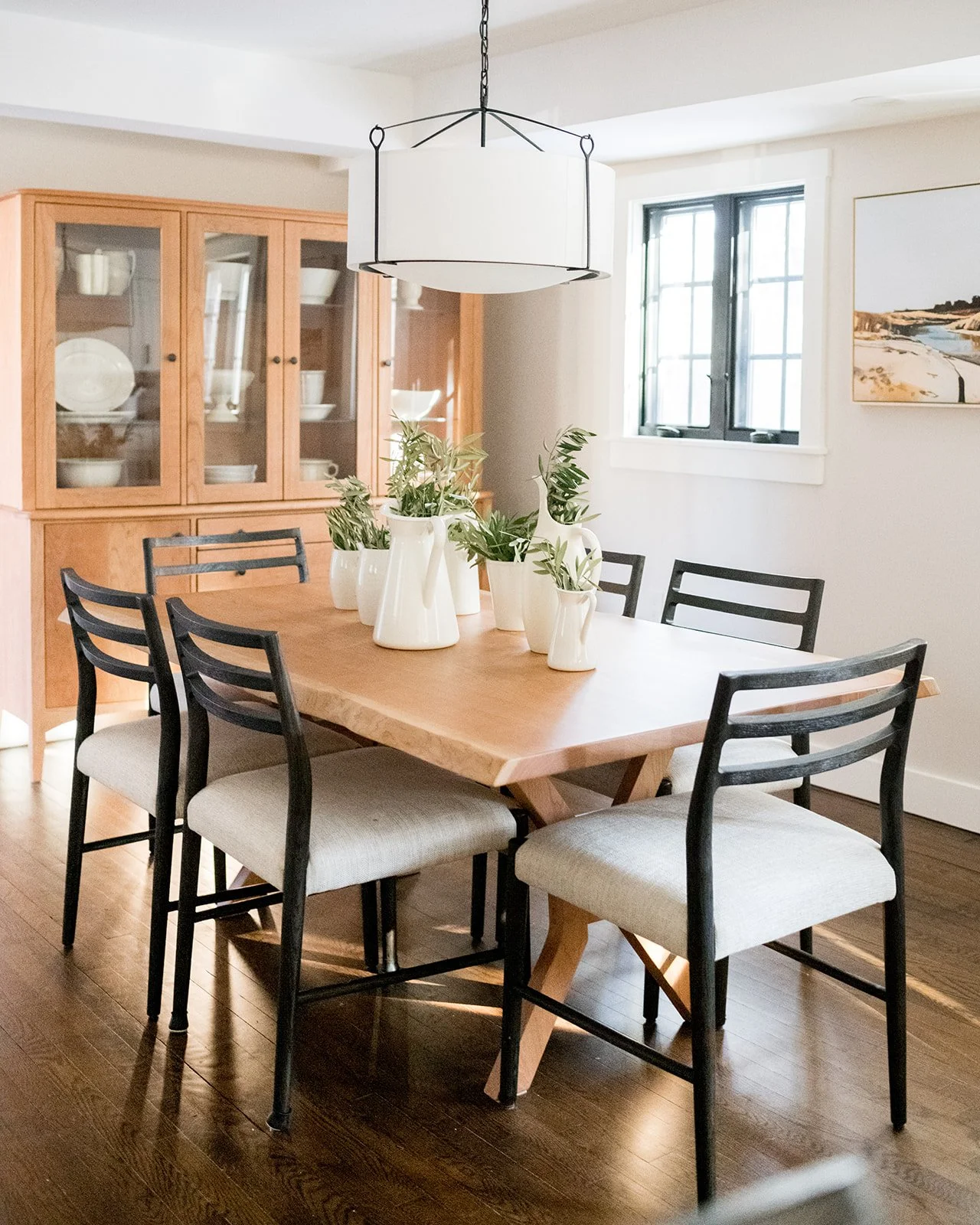 A dining room with a wooden table, surrounded by six black chairs with white cushions, and decorated with white vases holding green plants, illuminated by a modern white and black pendant light, with a wooden china cabinet and a window showing natural light in the background.