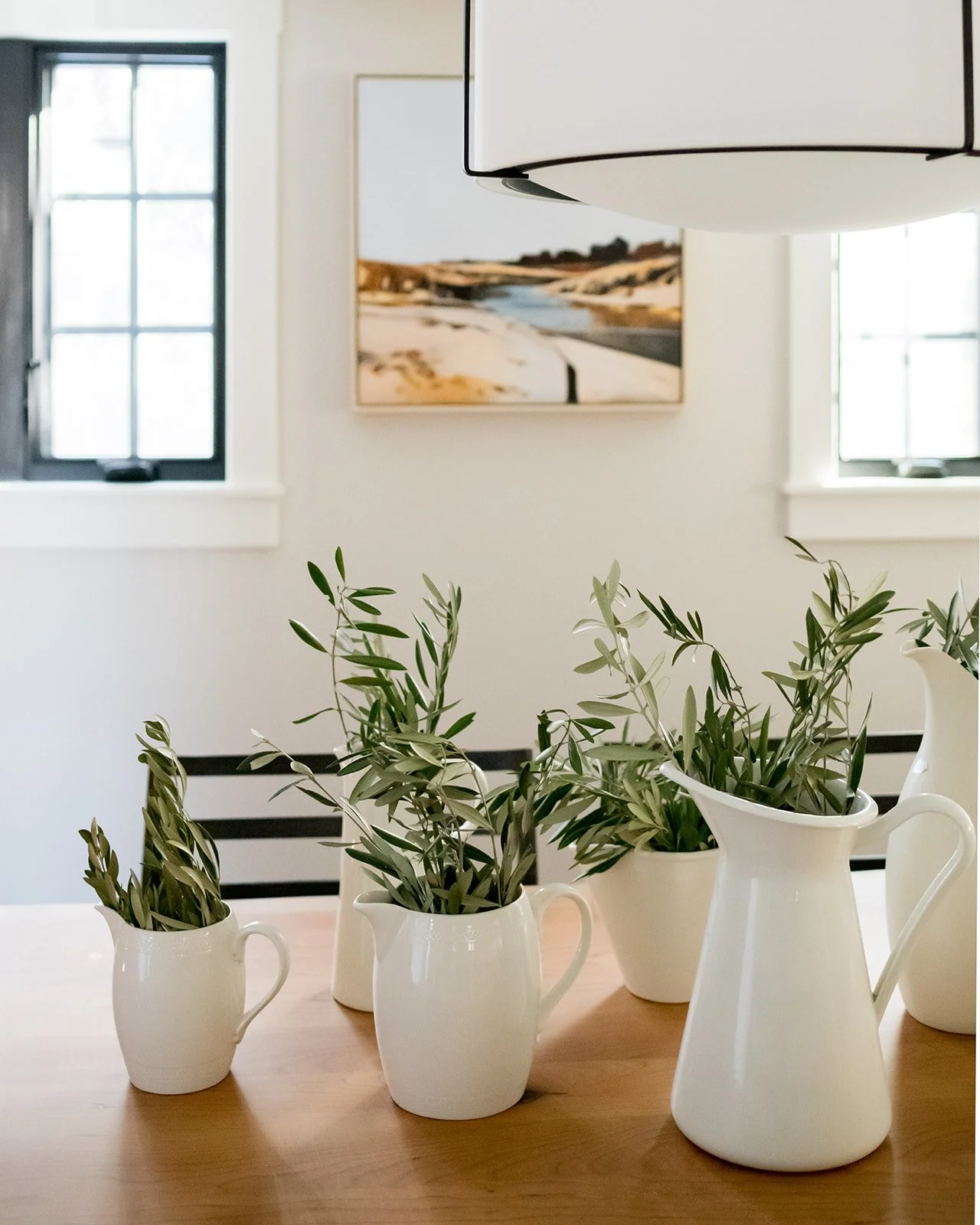 A wooden dining table with five white ceramic pitchers, each containing greenery. Two windows with black frames and a framed landscape painting are in the background, along with a white ceiling light.