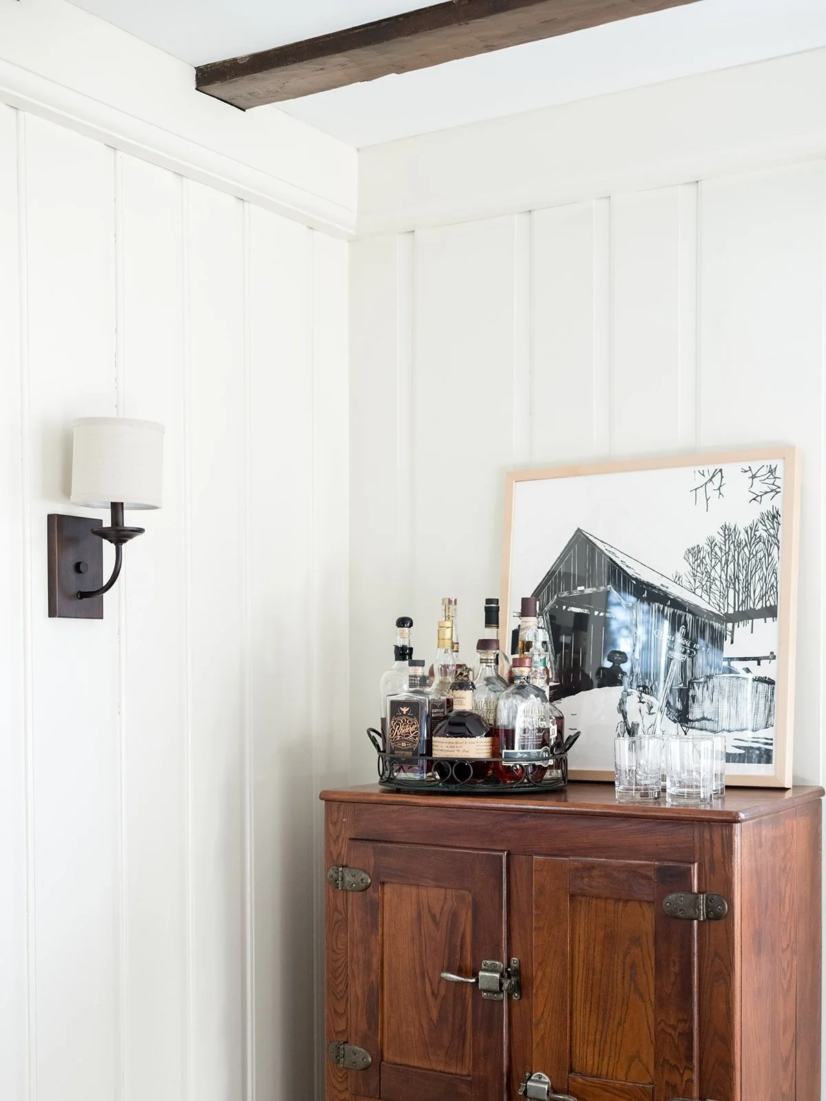 A wooden cabinet with bottles of liquor, a glass, and a framed black-and-white photo of a barn leaning against the wall in a room with white paneled walls and a wall sconce.