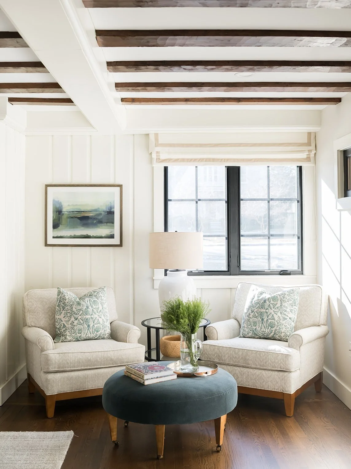 A cozy living room corner with two beige armchairs, a black side table with a white lamp, a round tufted ottoman with books and a plant, and a window with sunlight