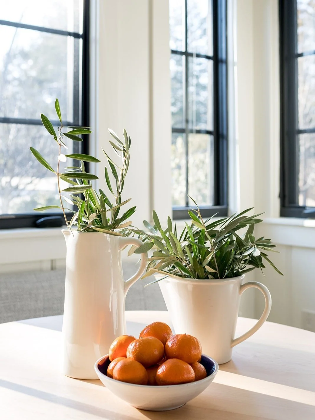Two white ceramic pitchers with green leafy plants, and a bowl of small round oranges on a white table near a window.