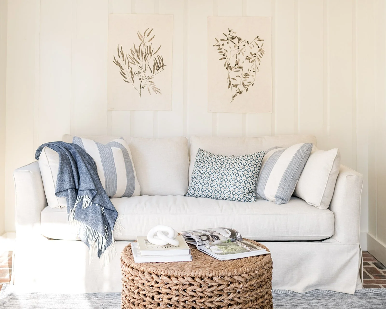A white couch with three pillows. One indigo-patterned pillow in the middle and two white pillows with blue stripes on each end. A blue throw blanket drapes over the left arm. Behind the couch are two botanical art prints on a white-paneled wall. In front of the couch is a woven round ottoman with an open magazine, a white decorative knot, and a plant catalog.