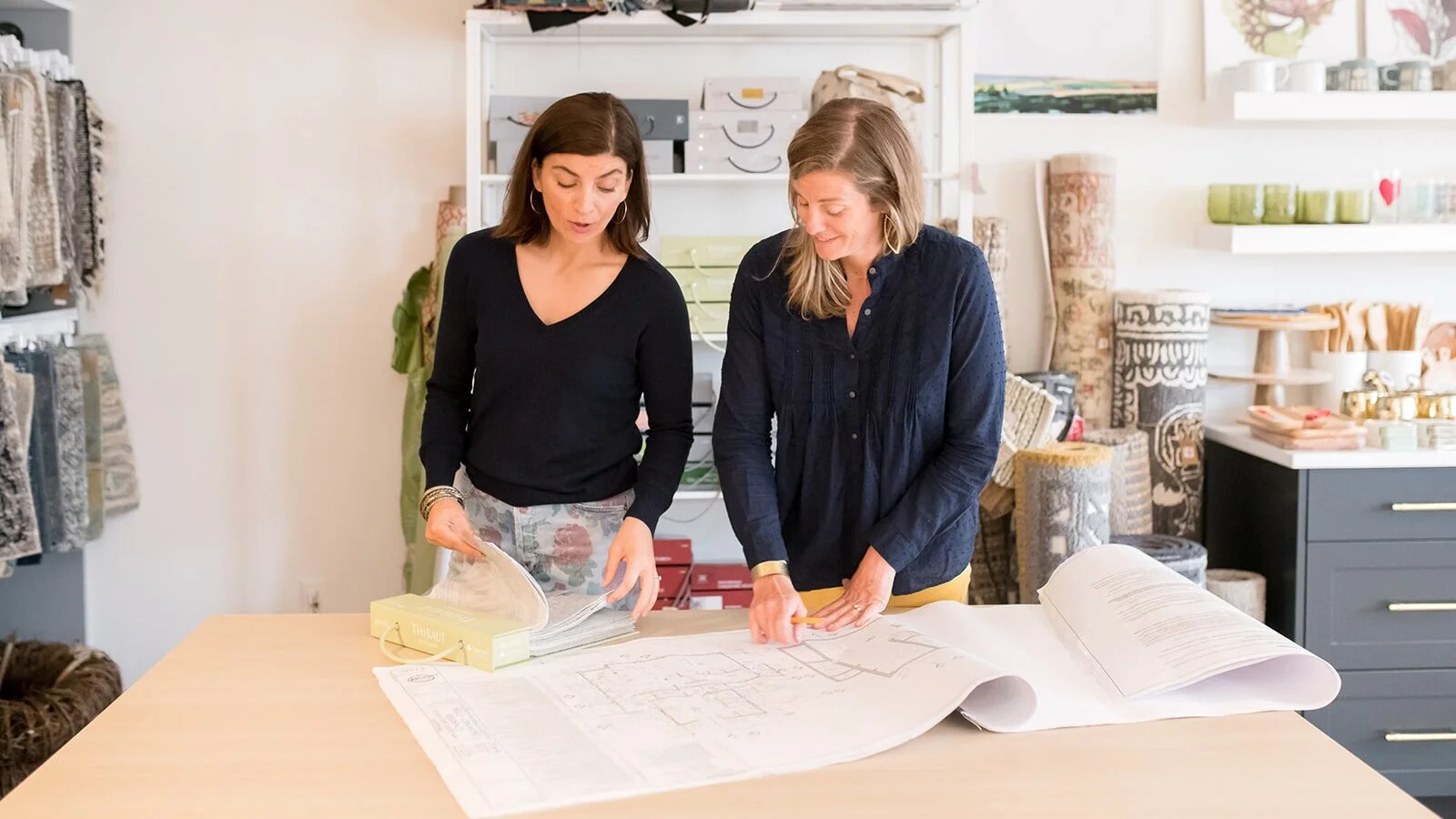 Two women look at a large architectural blueprint on a table in a home decor or fabric store.