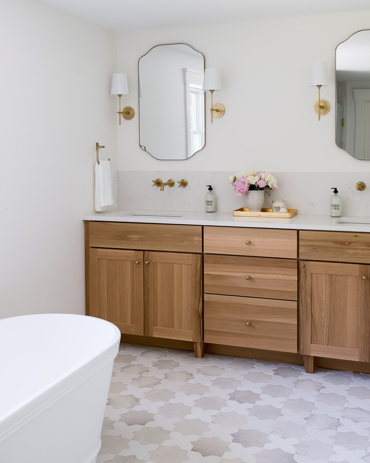 Bathroom with wooden vanity, white marble countertop, two oval mirrors, brass fixtures, wall-mounted lamps, pink and white flowers, and patterned tile floor.