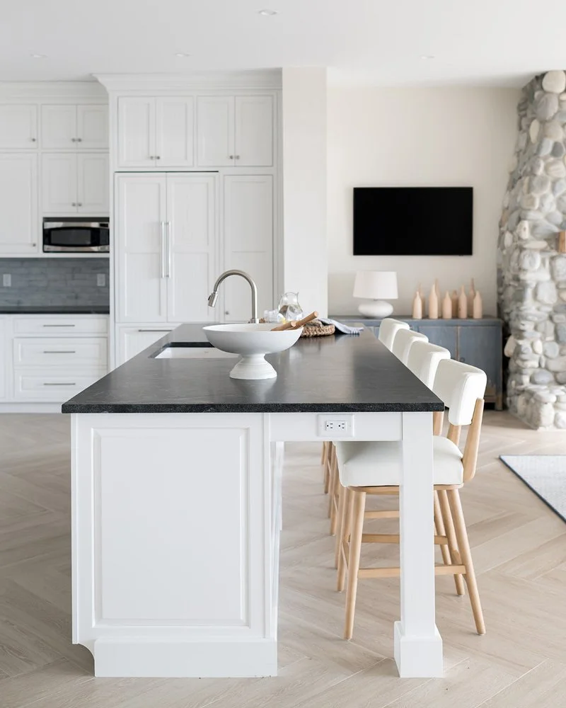 Modern kitchen with white cabinets, black countertop island with a bowl, barstools, and a living space in the background with a TV and stone fireplace.