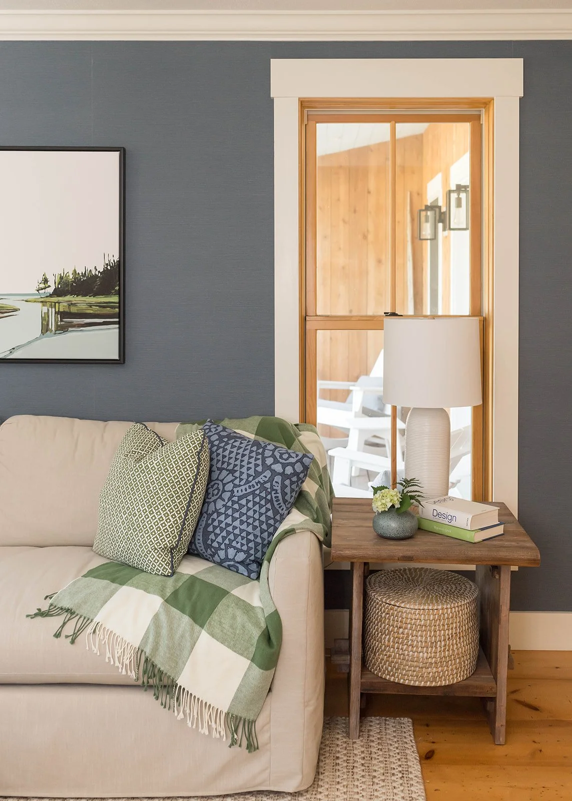 Living room with beige sofa, patterned pillows, green and white throw blanket, wooden side table with a vase of flowers, books, and a white lamp, blue wall, framed landscape artwork, and a wooden door with a view outside.