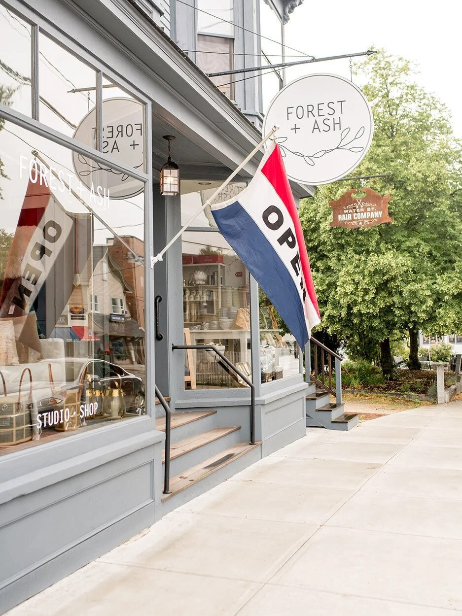 Exterior of a shop named Forest + Ash with large glass windows, a hanging lantern, and a vertical flag sign with an American flag design that reads "OPEN". There are stairs leading up to the entrance and a sidewalk in front.