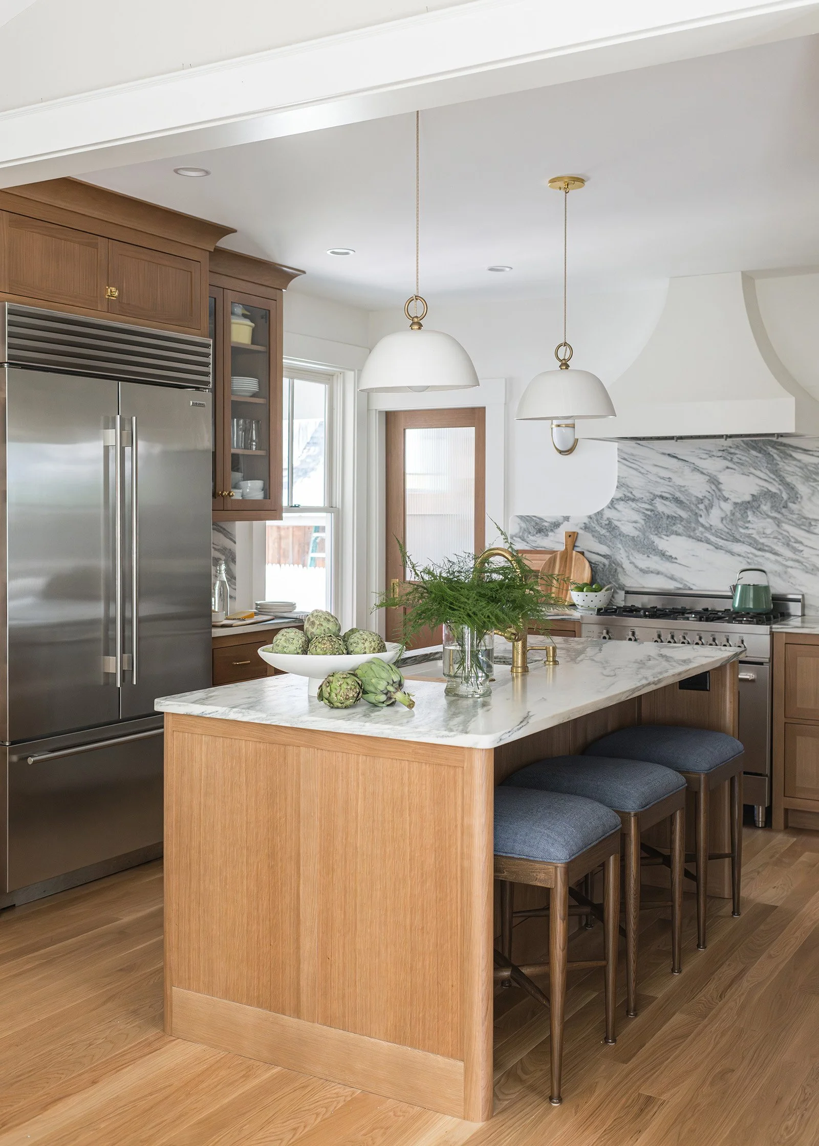 Modern kitchen with wooden cabinets, marble island counter, and pendant lighting. Artichokes are on the counter, and bar stools are at the island.