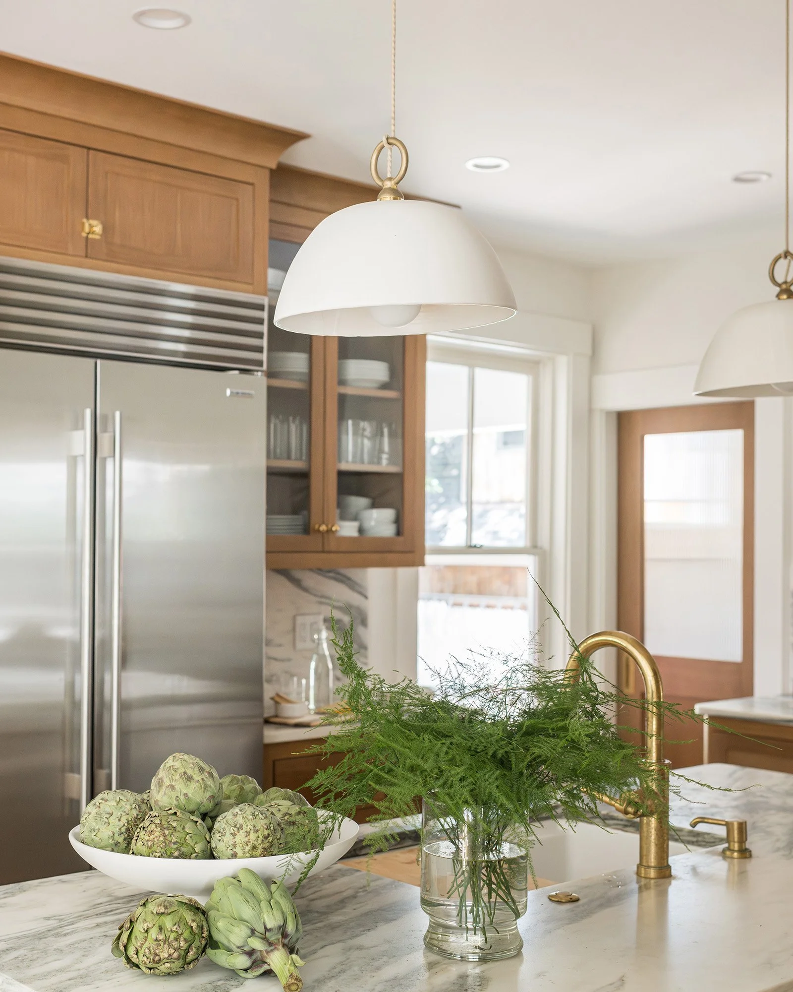 A kitchen countertop with artichokes in a white bowl, a bunch of greenery in a glass jar, and artichokes on the marble surface. Two white pendant lights hang from the ceiling, and there are wooden cabinets, a stainless steel refrigerator, and a window in the background.