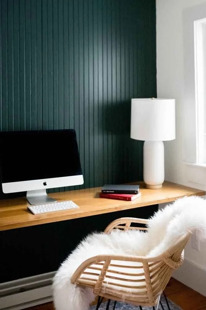 Home office desk with an Apple iMac computer, a white keyboard, a closed notebook, and a lamp on a wooden surface in a room with dark green beadboard wall and a white window.