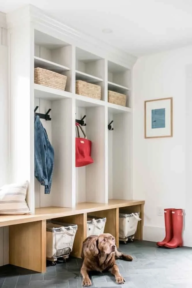 Entryway with built-in white cubbies, black hooks, and storage baskets, a bench with fabric bins underneath, red rain boots, a framed picture on the wall, and a brown dog lying on a dark tile floor.
