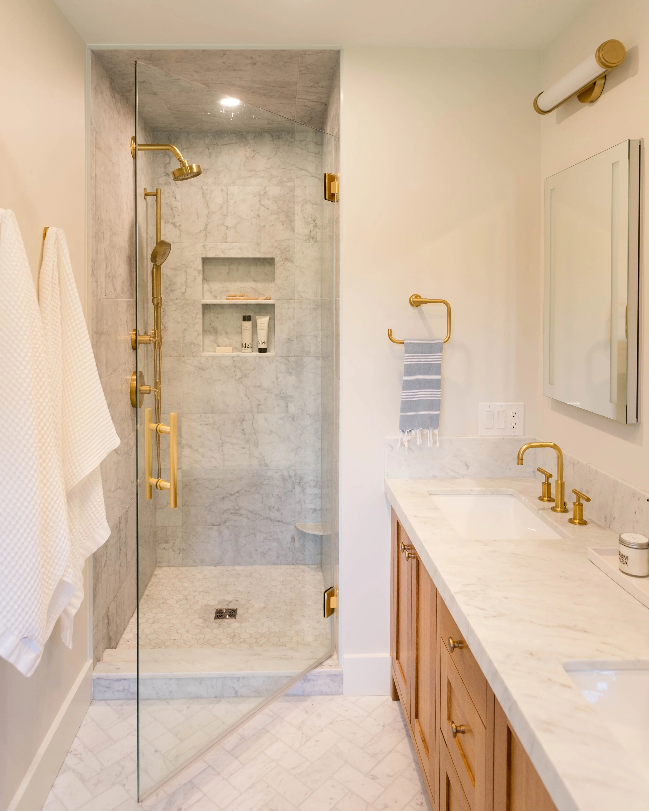 A bathroom with a marble shower enclosure and a wooden vanity with a marble countertop and gold fixtures.