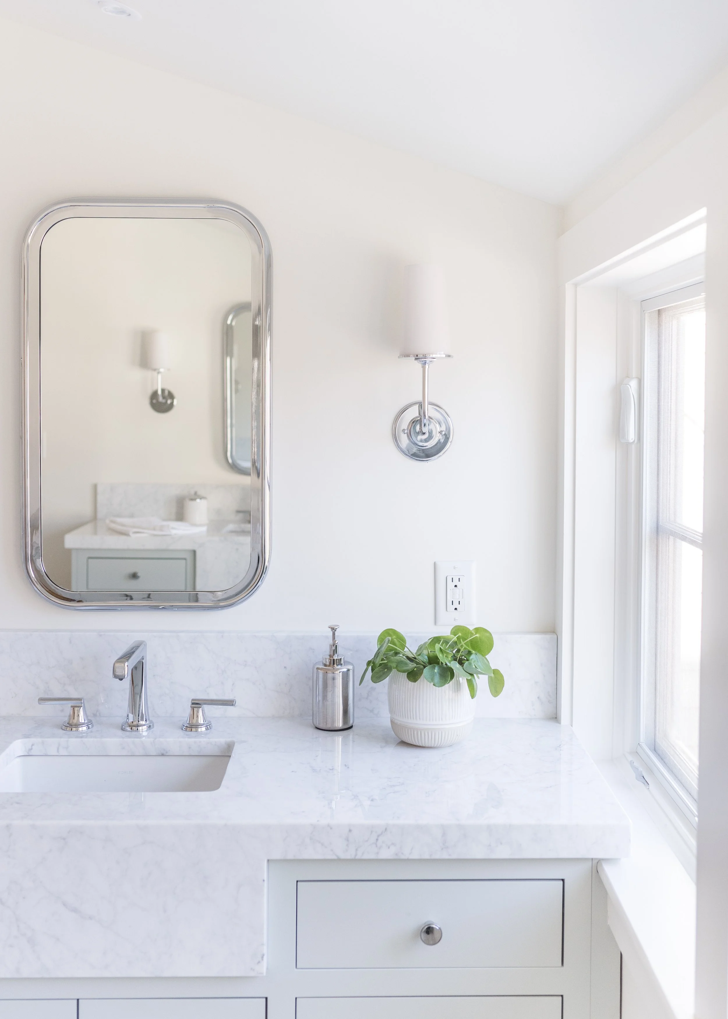 Bathroom vanity with a marble countertop, a built-in sink, and a silver faucet. There is a small potted green plant, a soap dispenser, and a mirror on the wall. A window provides natural light.