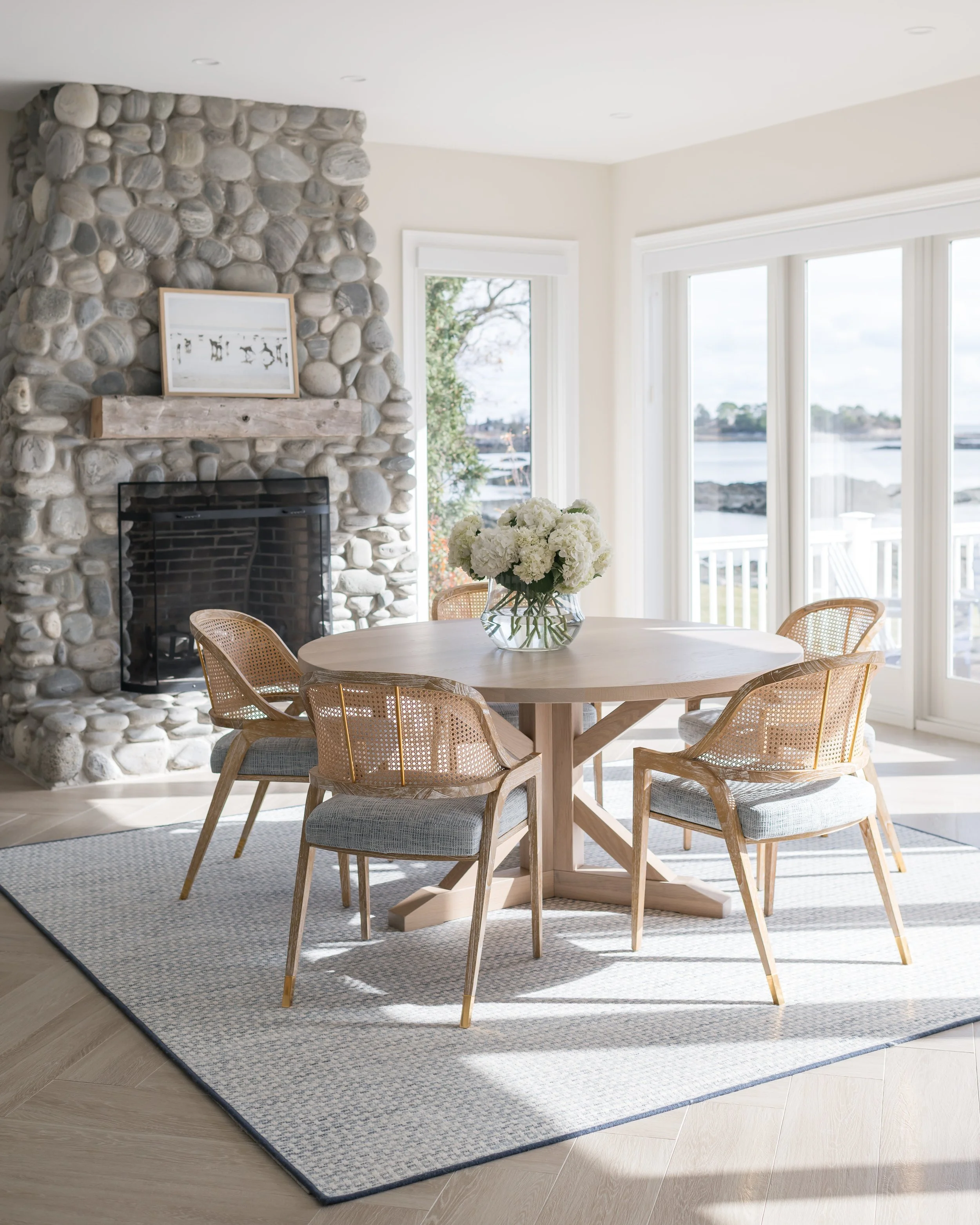 A dining area with a round wooden table, surrounded by six beige wicker chairs with cushions. A glass vase with white flowers is on the table. The space has large windows with a view of water and trees outside, and a stone fireplace with a framed photo on the mantel. There is a gray patterned rug on light wood flooring.