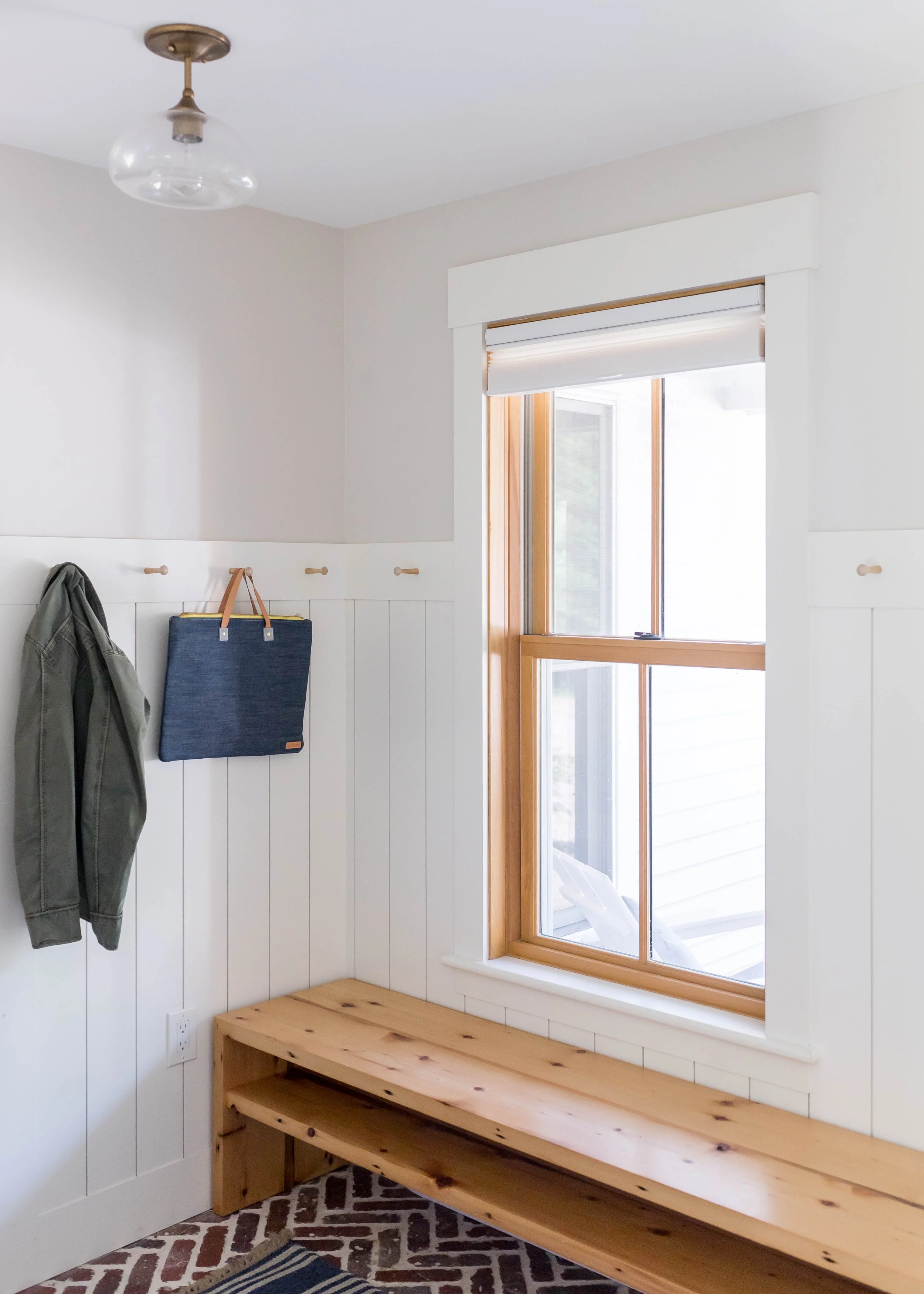 Interior of a bright room with white walls, a window with wooden trim, a wooden bench, and hooks on the wall, with a coat and tote bag hanging. There's a patterned rug on the floor.