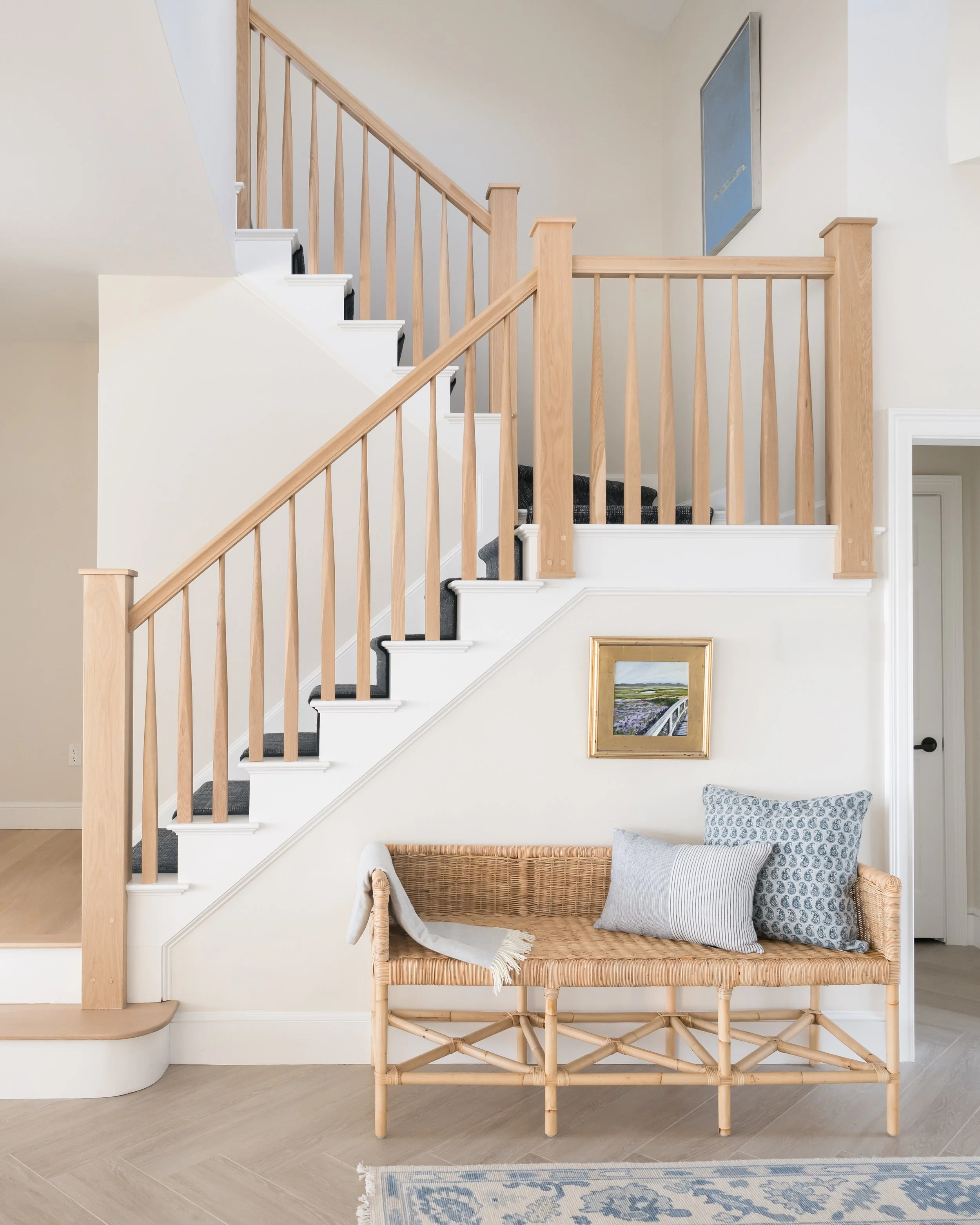 Interior view of a staircase with a natural wood railing and white risers, black treads, and a carpet runner. A woven bench with three decorative pillows is placed underneath the staircase, next to a framed landscape painting on the wall, and a blue and white patterned rug on the light wood floor.
