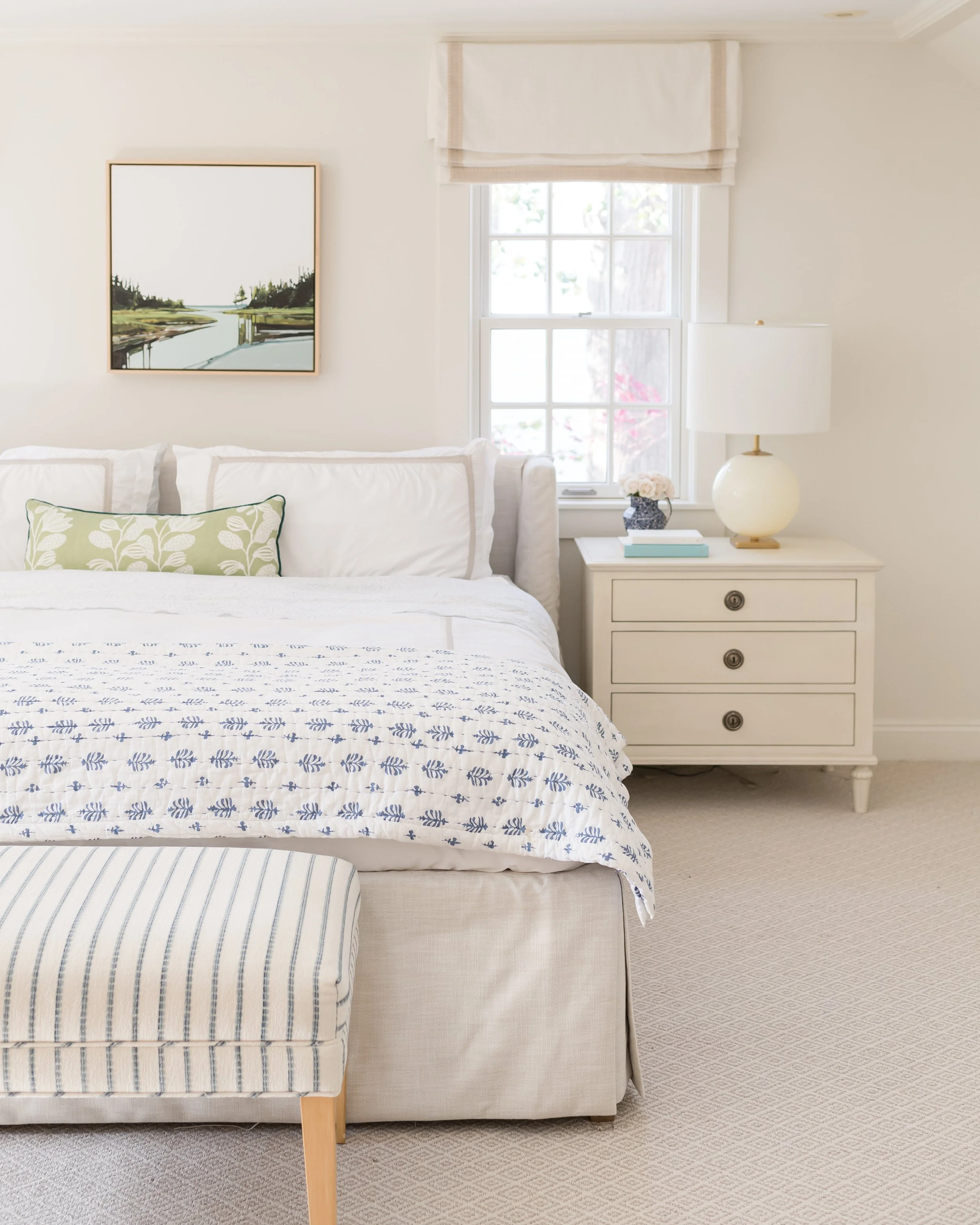 A bedroom with a white bed, a green and white decorative pillow, and a third striped pillow. A white nightstand with three drawers, a white lamp, books, and a small flower vase. A window with sunlight, a white curtain, and an outside view. A piece of framed artwork depicting a waterway with trees on each side hangs above the bed.