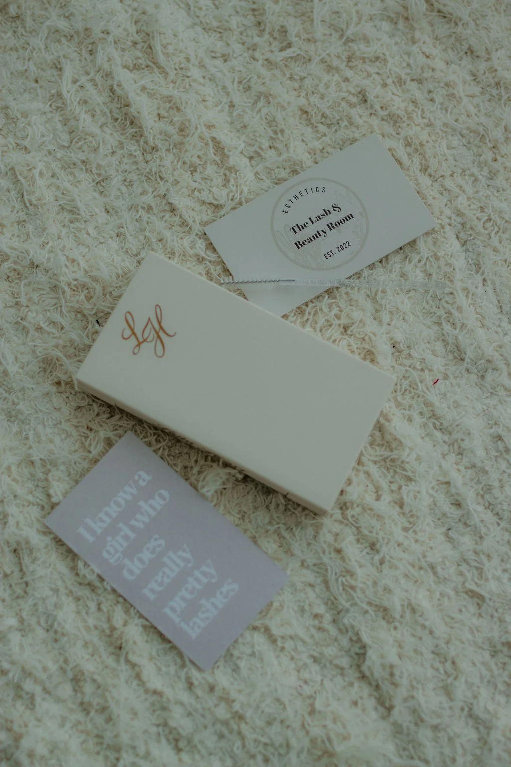 Business cards and a white box placed on a fluffy cream-colored surface. One card reads "The Lash & Beauty Room," another has the initials "LBR" in red, and a third card says "Quality aloe vera really pastes."