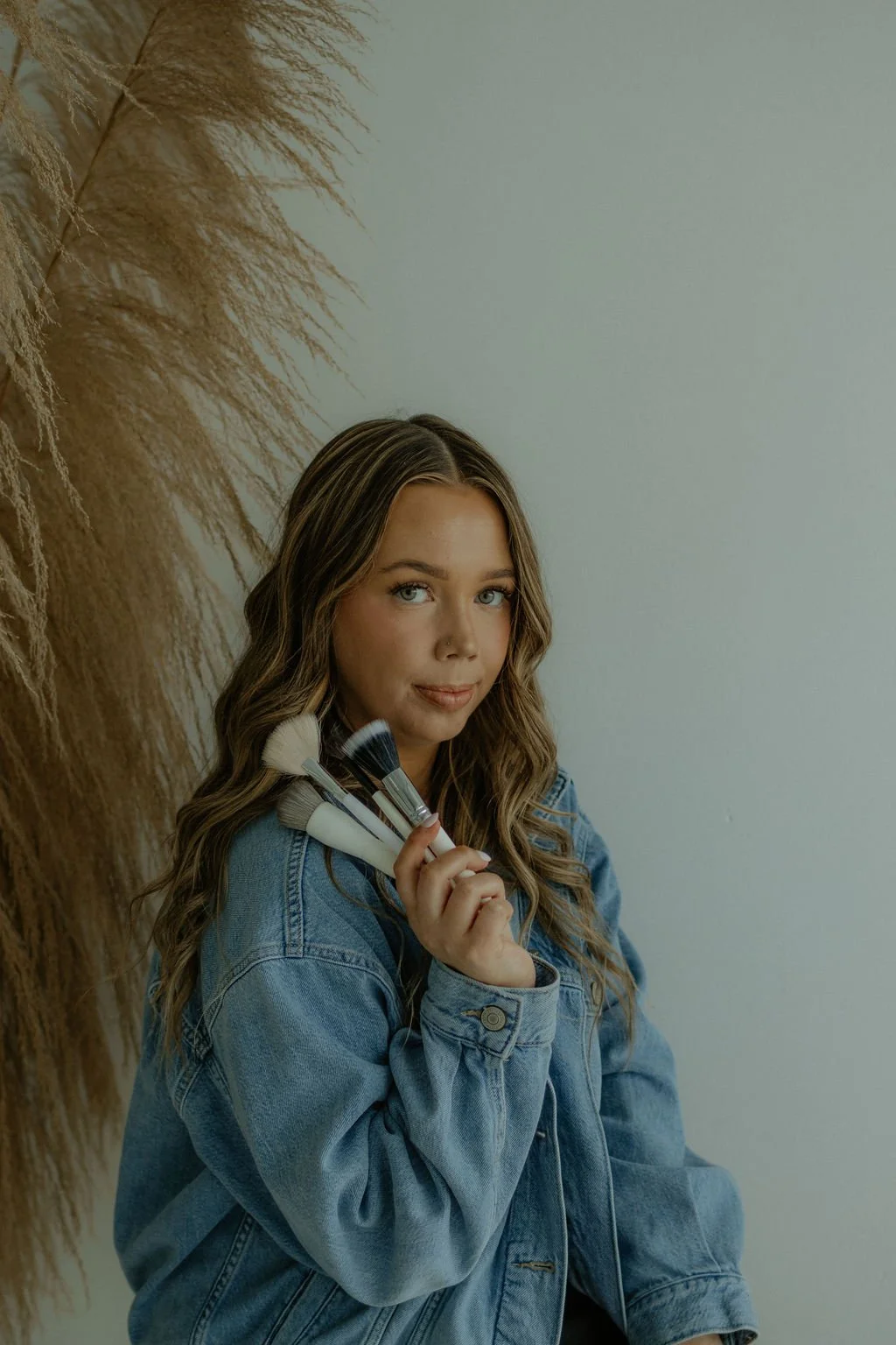Young woman with wavy brown hair, wearing a denim jacket, holding makeup brushes, and posing against a plain wall with dried pampas grass.