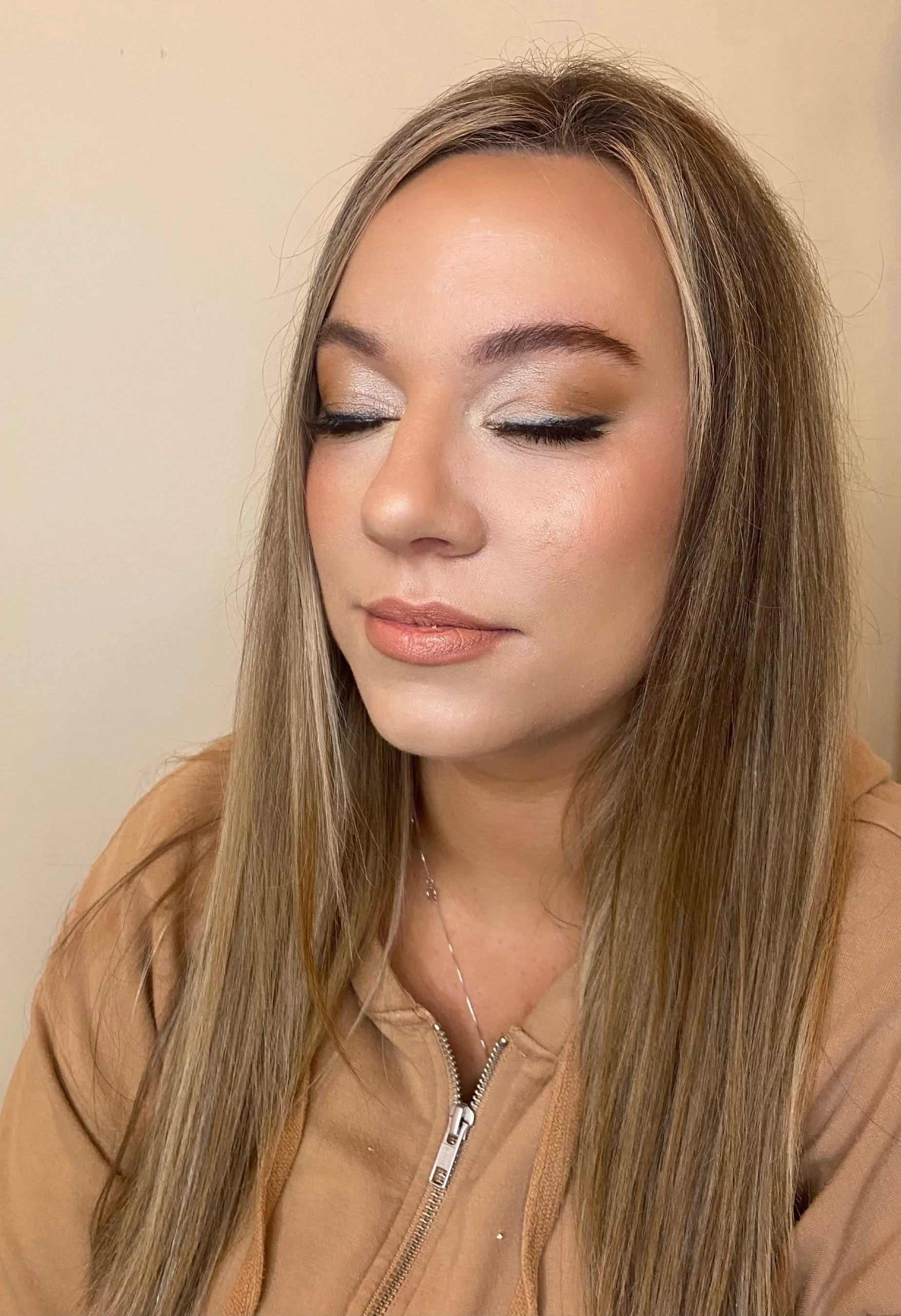 A young woman with long, straight, light brown hair and fair skin closed her eyes. She is wearing a tan zip-up jacket and a delicate silver necklace.