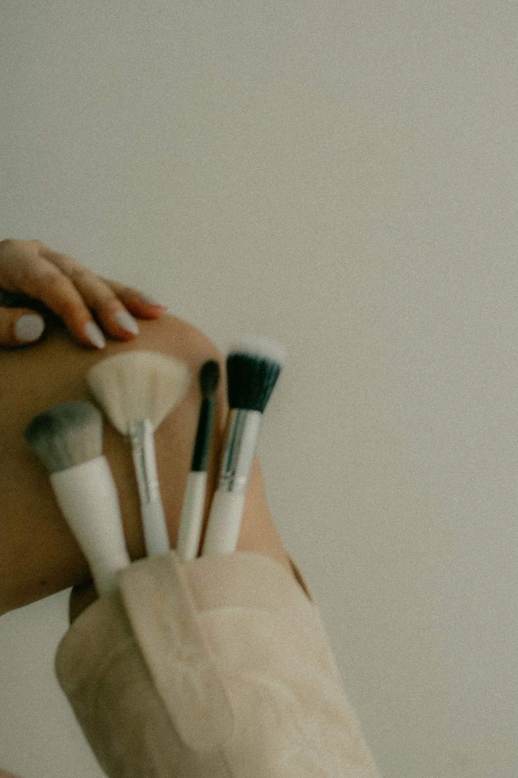 A person holding a shopping bag with three makeup brushes sticking out, touching their leg which has a hand resting on it, against a plain ceiling background.