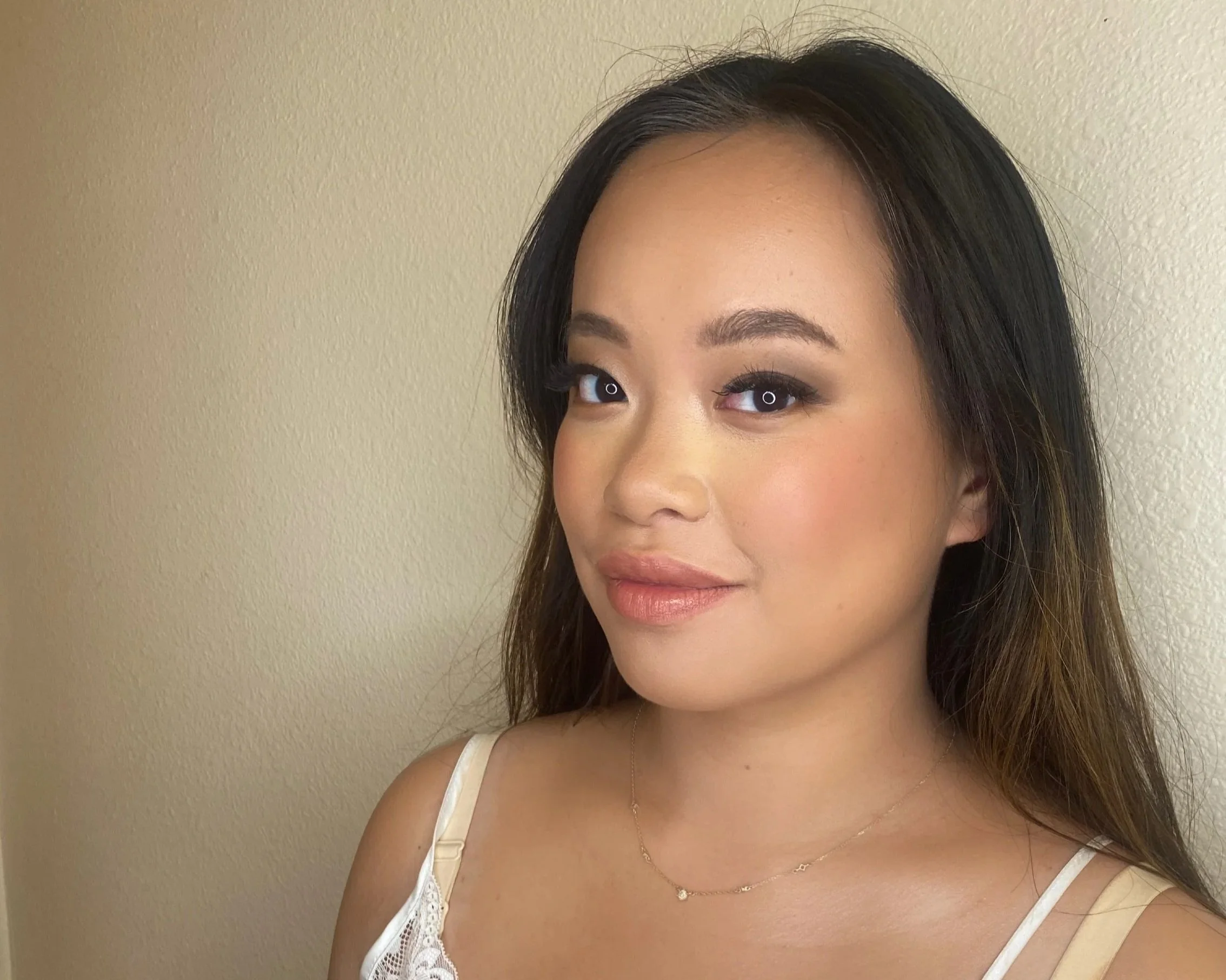 Close-up of a young woman's face with makeup, wearing a white lace top and a necklace with a small pendant, standing against a plain beige wall.