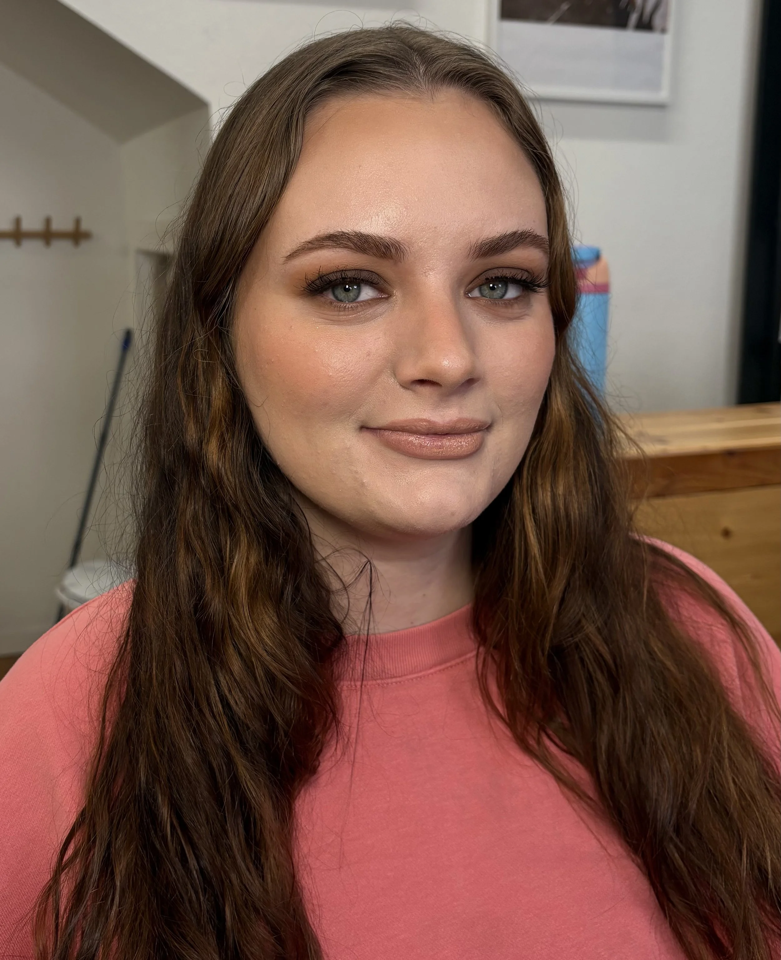 A woman with long brown hair and blue eyes looking at the camera, wearing a pink top, in an indoor setting.