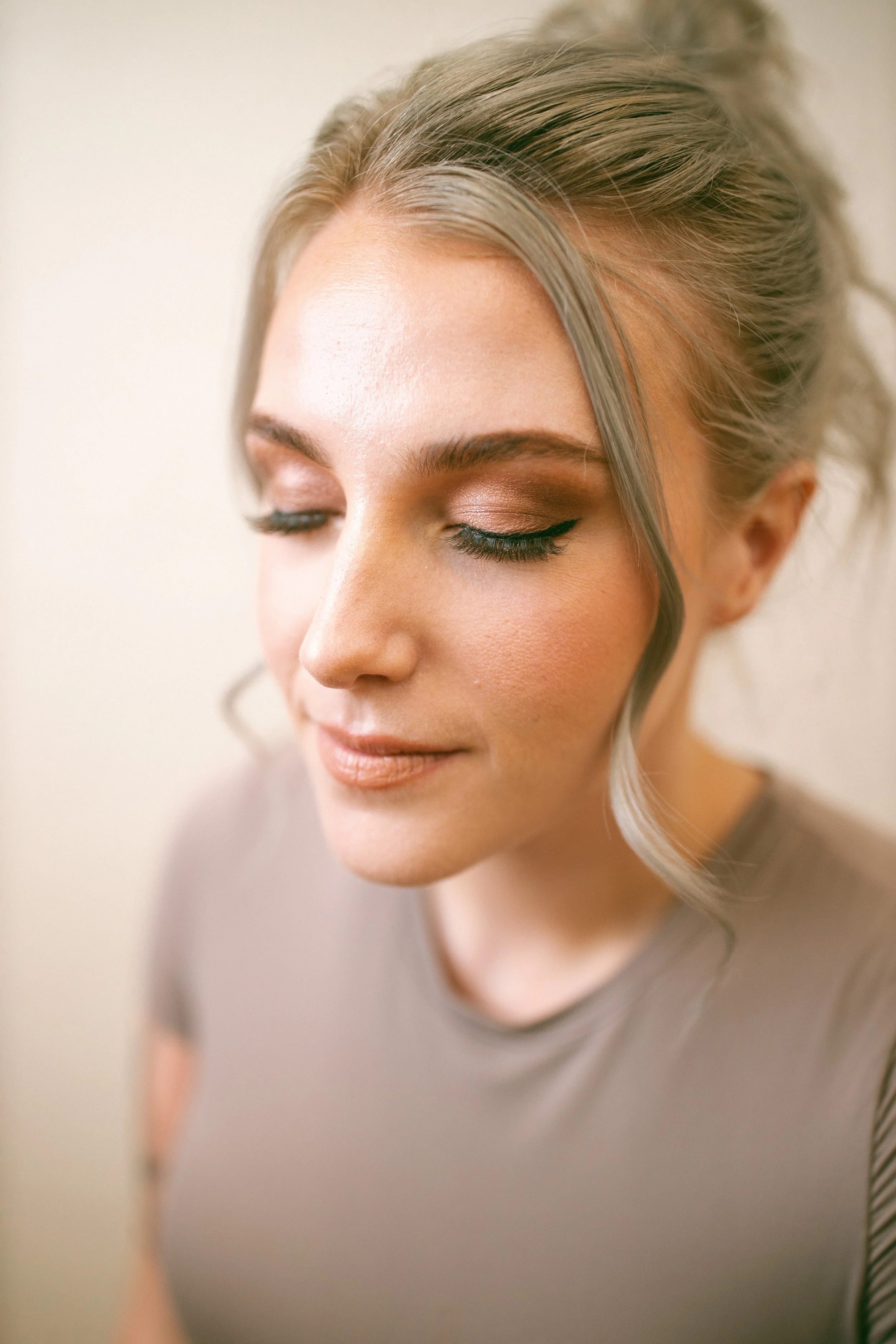 Close-up of a woman with blonde hair styled in an elegant updo, wearing soft makeup with accentuated eye makeup, a winged eyeliner, and nude lipstick.