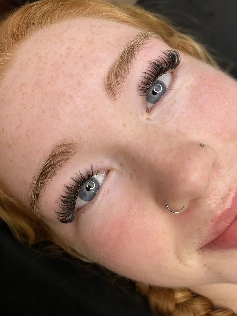 Close-up of a woman's face showing her blue eyes, eyebrow eyelashes, freckles, and facial piercings including a nose ring and a small nose stud.