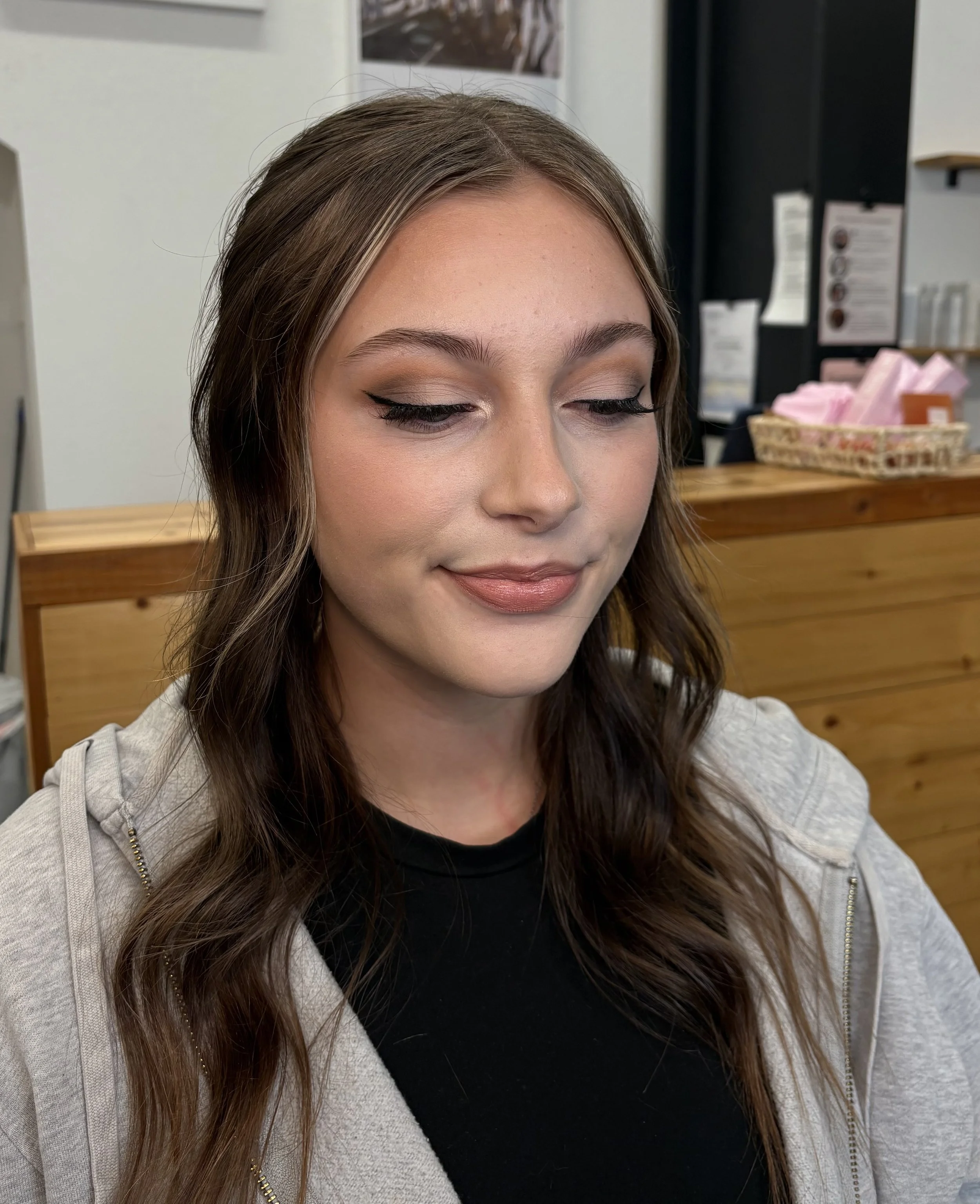 A woman with long wavy brown hair, wearing makeup including eyeliner and lipstick, faces downward with closed eyes, smiling slightly, in an indoor setting with a wooden furniture piece and various posters and items in the background.