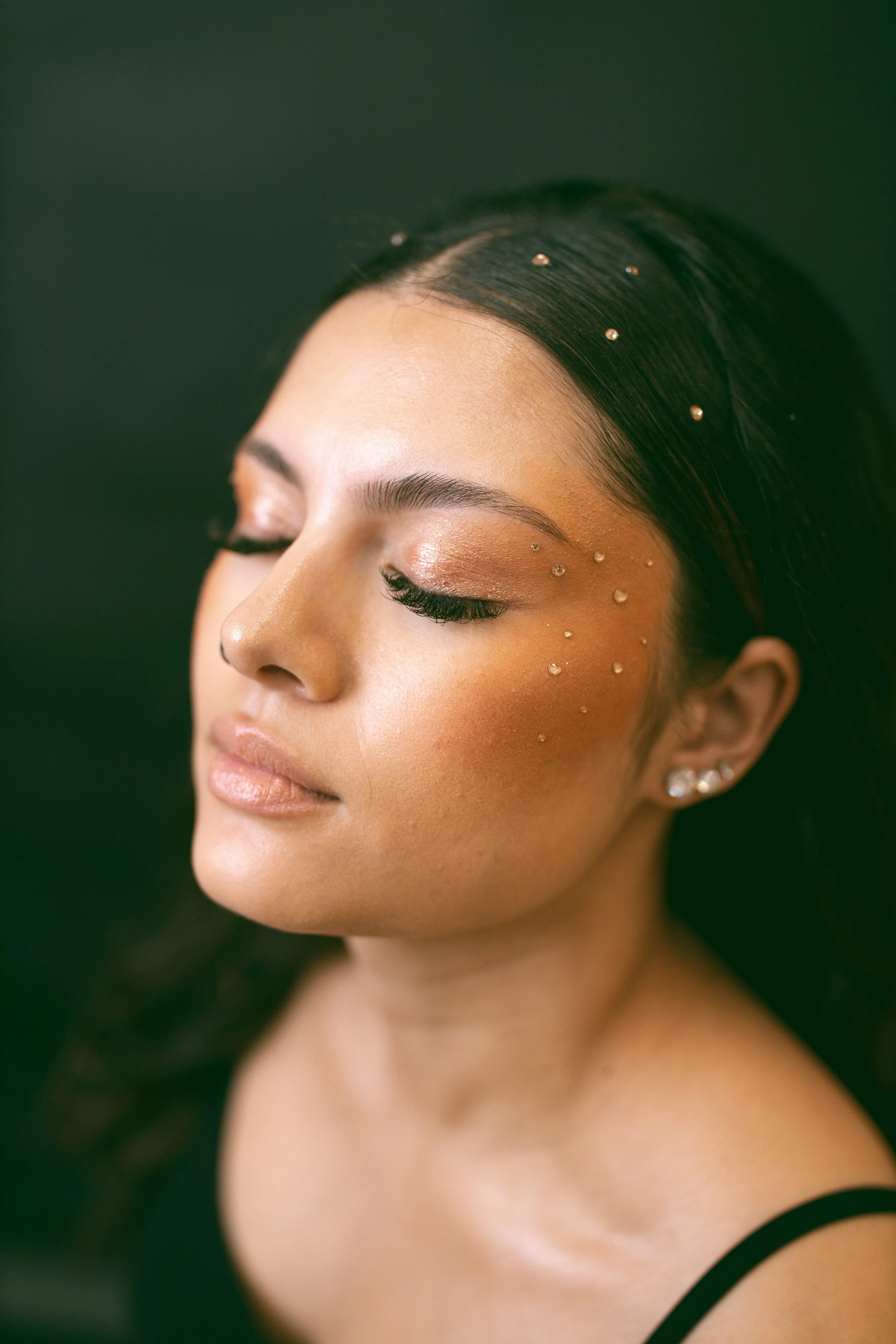 A close-up of a woman's face with closed eyes, adorned with small decorative gems near her eye and ear, with smooth skin and dark hair against a dark green background.