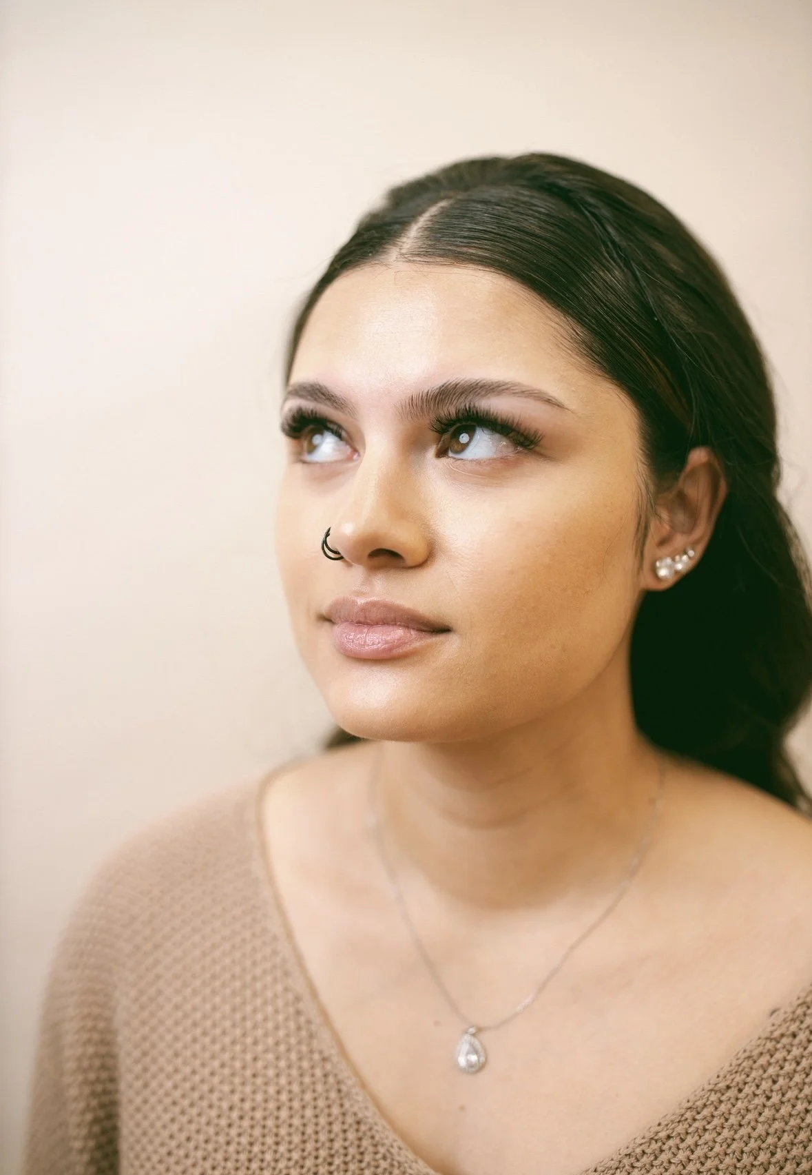 A young woman with dark hair, light makeup, and multiple earrings looking upwards and to the side, wearing a beige sweater and a silver necklace.