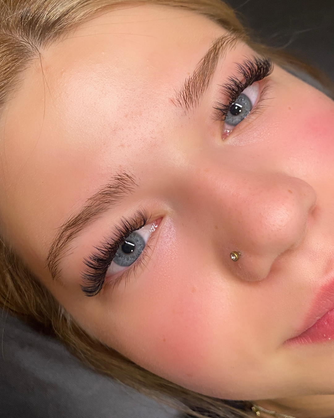 Close-up of a woman's face with light makeup, blue eyes, long eyelashes, well-groomed eyebrows, and a nose piercing.