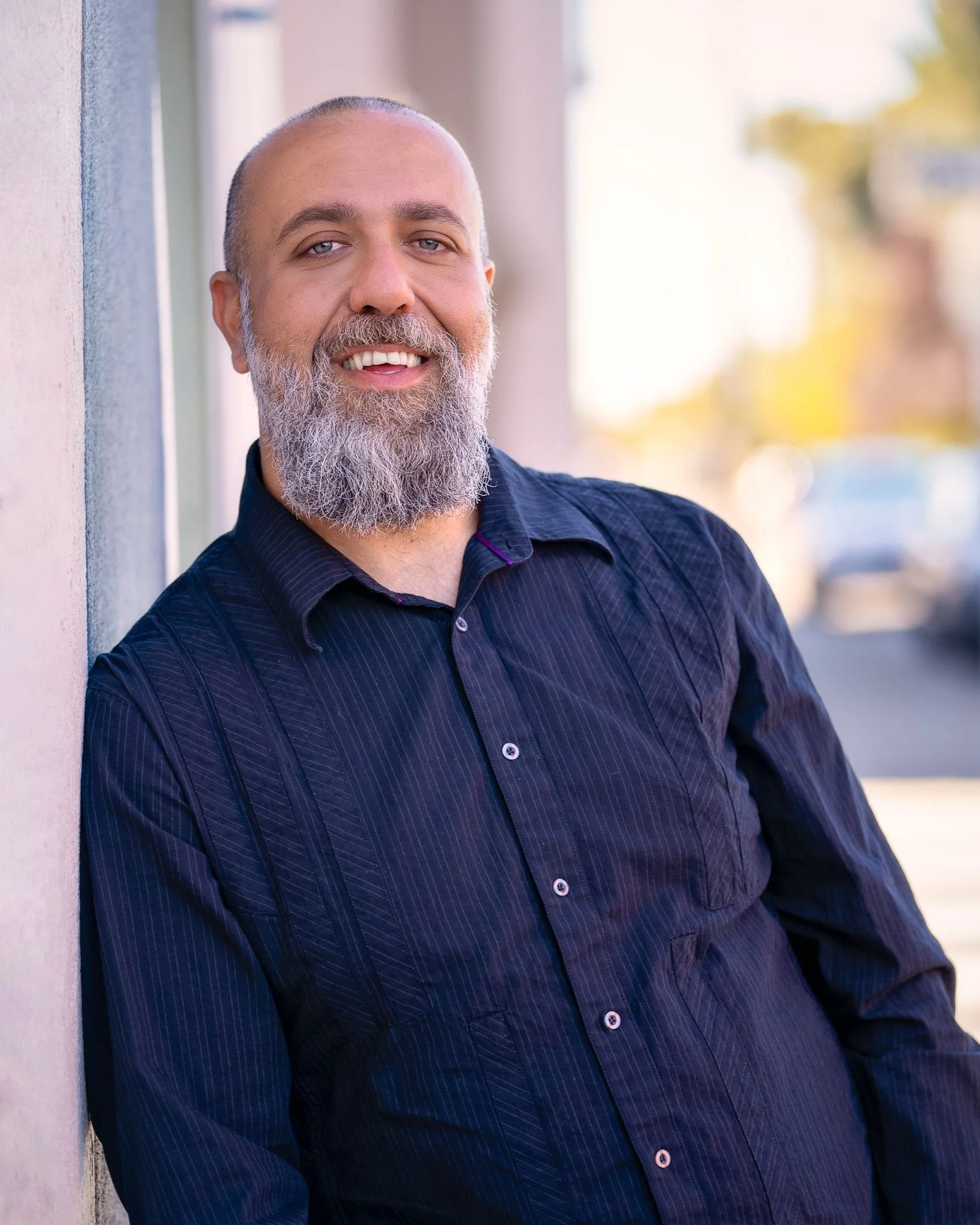 A middle-aged man with a beard and a bald head, smiling and leaning against a wall outdoors during daytime.