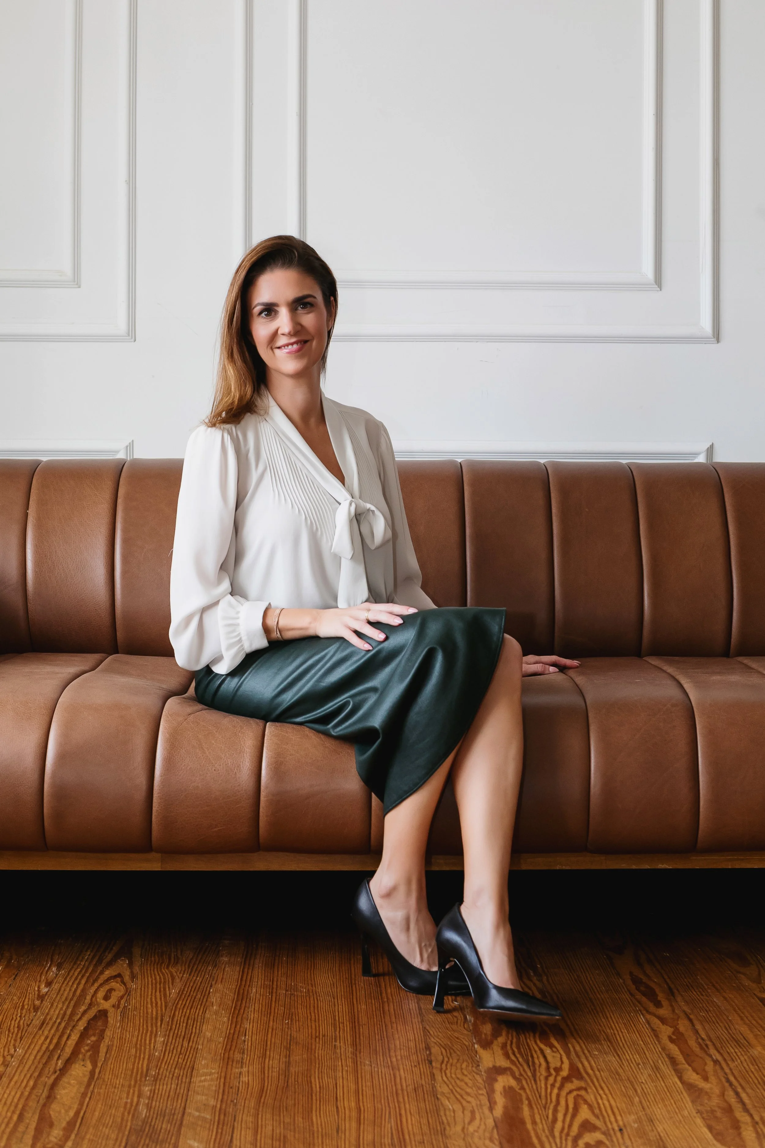 A woman with brown hair sitting on a brown leather bench against a white wall with decorative molding, wearing a white blouse, green skirt, and black high heels.