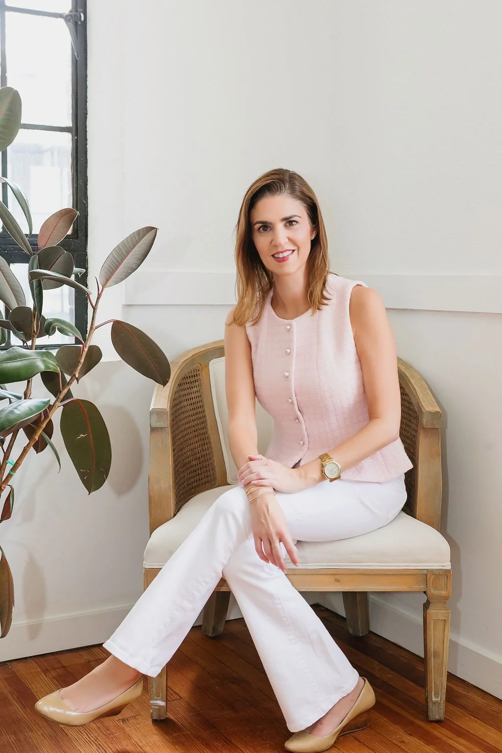 A woman sitting on a wooden bench in a room with white walls, a large plant, and a window with black framing. She is smiling and wearing a pink sleeveless top, white pants, beige heels, and a gold watch.