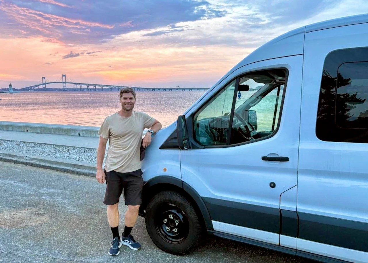 A man standing next to a light blue van near a body of water during sunset, with a bridge in the background.