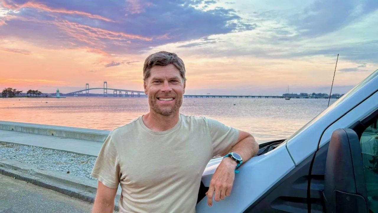 A man standing next to a vehicle on a waterfront at sunset, with a bridge in the background.