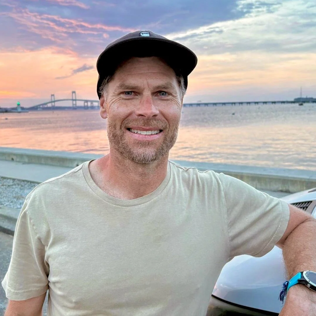 A man wearing a beige t-shirt and a hat, smiling at the camera, stands by a waterfront during sunset with a bridge in the background.